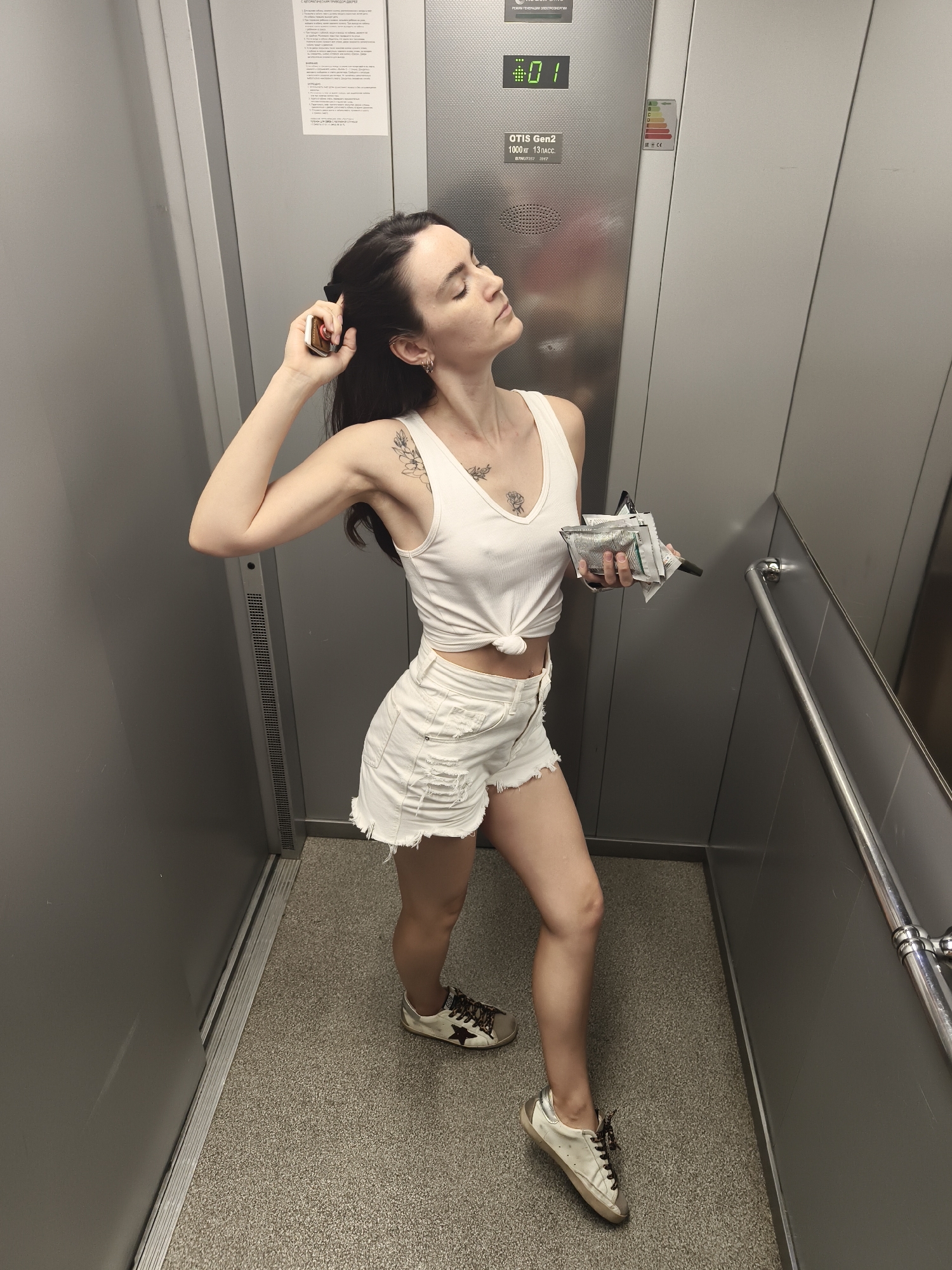 The image features a young woman posing inside a modern elevator in Tyumen, Russia. She has dark hair and is dressed in a white ribbed tank top, tied at the waist, and white distressed denim shorts. She wears off-white sneakers with a star emblem. Visible tattoos include floral designs on her left shoulder and chest. The woman stands with her head tilted back, eyes closed, and a relaxed expression, her right hand near her hair, holding a small metallic object. In her left hand, she holds several small, silver-foil packets and a dark, slender item.
The elevator interior features metallic grey walls and a dark, patterned carpeted floor. A silver handrail is visible on the right side. On the elevator panel, a digital display shows "01," indicating the first floor. Below this, text reads "OTIS Gen2," "1000 кг 13 пасс" (1000 kg, 13 passengers), and "8784T257 2017." An energy efficiency label prominently displays 'A' as the highest rating. A large white information sheet with text in Cyrillic script is posted above the panel. The lighting suggests an indoor setting, possibly daytime.