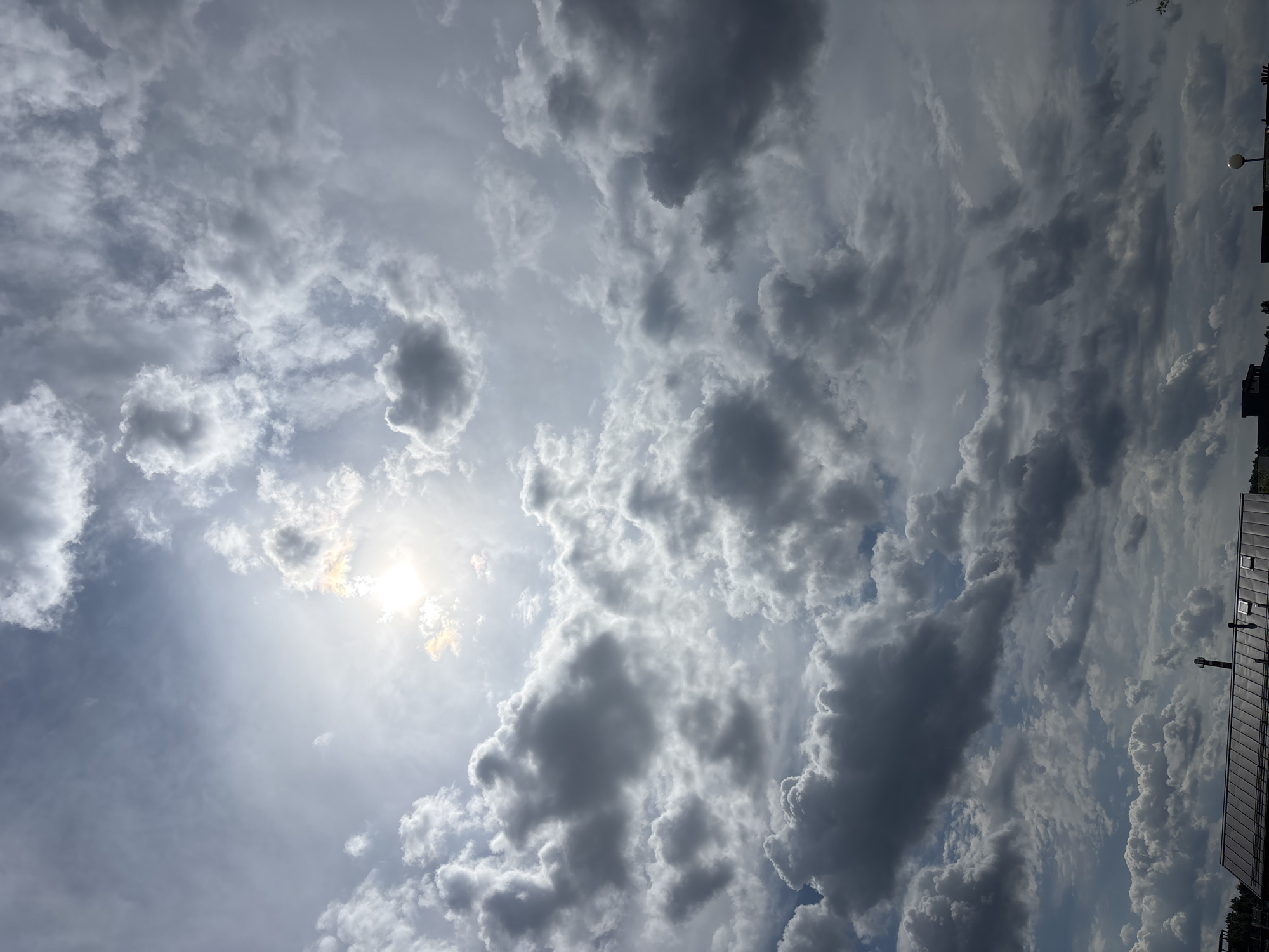 The image depicts a dynamic sky over Zvenigorod, Russia, characterized by a dramatic interplay of sunlight and clouds. The sun, partially obscured by luminous clouds, casts a bright glow on the upper left quadrant of the scene, creating a halo effect and highlighting the texture of the surrounding cumulus and stratocumulus formations. The clouds vary in color and density, ranging from bright white and grey to darker, more ominous shades, suggesting a weather system in transition. The lower portion of the image reveals the rooftops of buildings, appearing to be in a residential or urban area, with some visible architectural details like chimneys and skylights. The overall impression is one of a powerful, natural spectacle unfolding above the city.