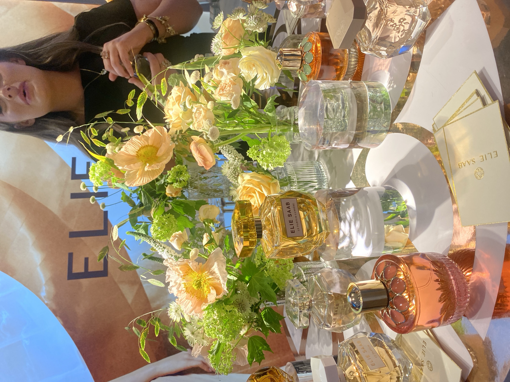 This media file captures an elegant display of Elie Saab fragrances and floral arrangements, likely at a product launch or promotional event in London.
In the upper left, a woman with long, dark hair and a black top is partially visible. Her face is in profile, and she wears several bracelets and rings on her hands, which are positioned near a perfume bottle. Her expression is neutral.
The foreground and center are dominated by a lush floral arrangement featuring various light yellow and peach-toned roses, delicate poppies with prominent yellow centers, and an assortment of green foliage and small white filler flowers. These are displayed in clear glass vases.
Surrounding the flowers are numerous Elie Saab perfume bottles of different shapes, sizes, and colors. Some are clear, others are in shades of golden amber, and one notable bottle is a reflective rose gold with intricate pink decorative elements around its cap. These bottles are arranged on a reflective surface, possibly a mirrored table, which features white and gold geometric patterns. Several small white cards with gold edges, bearing the "ELIE SAAB" logo, are also scattered on the table.
In the background, a large display or banner is partially visible, showing the brand name "ELIE SAAB" in large, dark letters on a light brown background. The lighting suggests a bright, indoor setting, possibly with natural light.
Visible text includes: "ELIE SAAB" on the large background banner, on the small cards, and prominently on multiple perfume bottles. Some bottles also bear "LE PARFUM" and one specifically states "LE PARFUM LUMIERE".