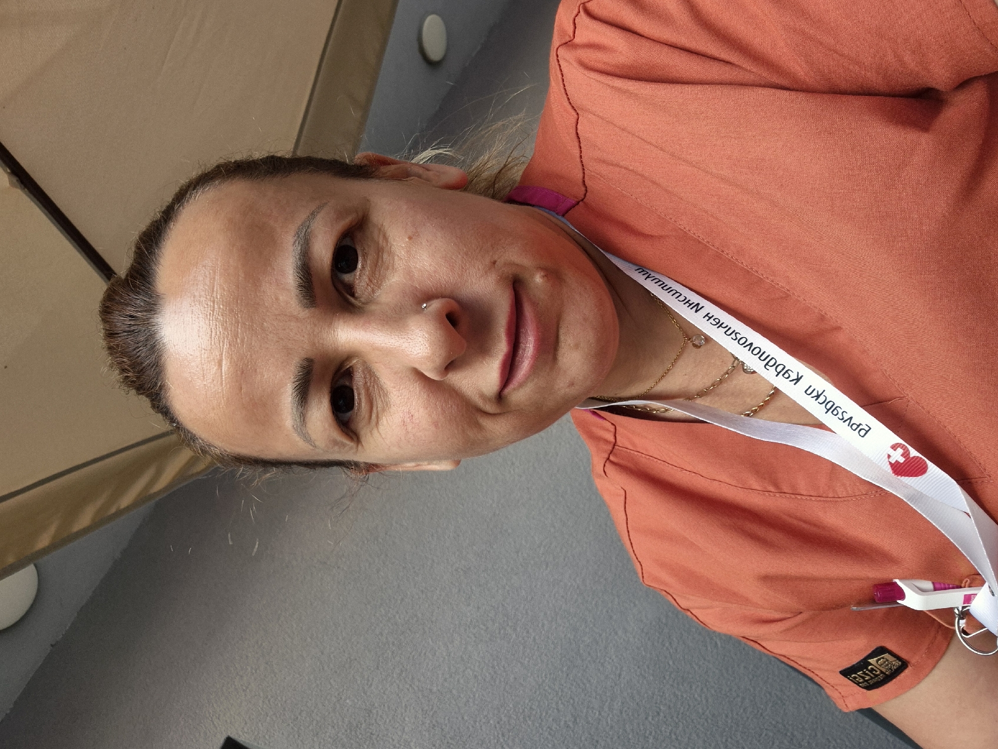 A medium shot photo shows a woman with light skin and brown hair pulled back. She is wearing an orange scrub top, and a white lanyard with a red heart and cross logo is visible around her neck. The background is a gray wall and an outdoor canopy. The woman has a nose ring and is smiling slightly. The image was taken outdoors in an urban environment.