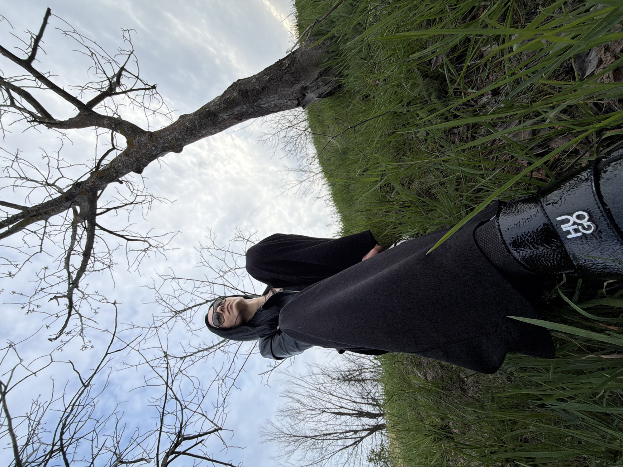 The image presents a low-angle view of a person lying in a grassy field beneath a bare tree. The person is dressed in black clothing, including a hooded garment, and sunglasses. Their head is positioned near the tree's branches, which reach towards a sky with light clouds. The lower portion of the image shows green grass, some of which obscures a black bag. The location is Pleven, Bulgaria.