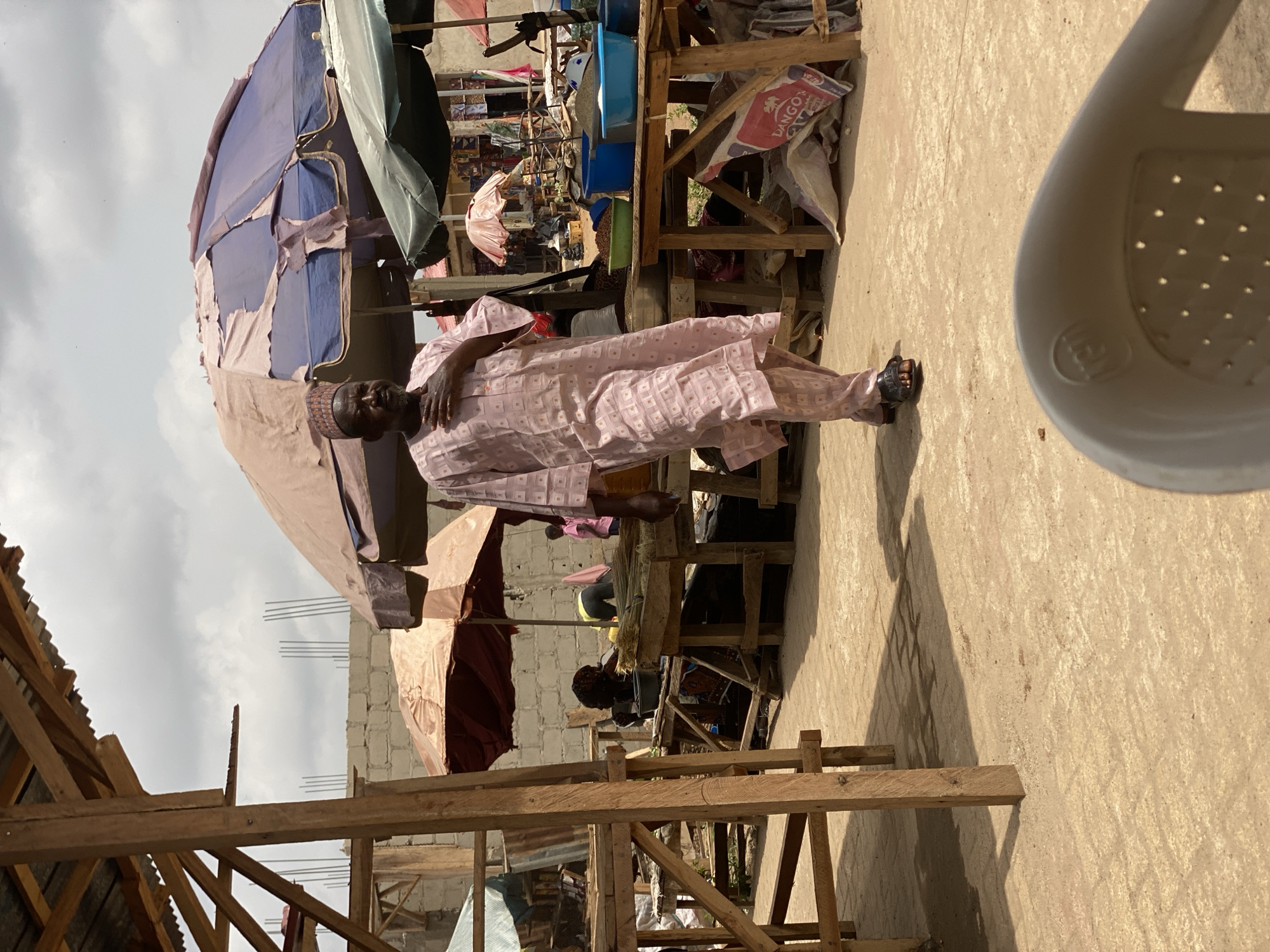 The image shows an outdoor scene in what appears to be a marketplace. An elderly man in a pink patterned robe and cap stands facing the camera. A dilapidated market stall with blue and beige tarps provides shade. Other stalls and merchandise are visible in the background. Wooden structures, possibly parts of stalls or buildings under construction, are scattered around. The ground is a beige color, possibly concrete or packed dirt. A portion of a white plastic chair is visible in the upper right corner. There are people and merchandise within the market, with a building made of cinder blocks and rebar visible in the background. The location is the city of Kafanchan B, Nigeria.