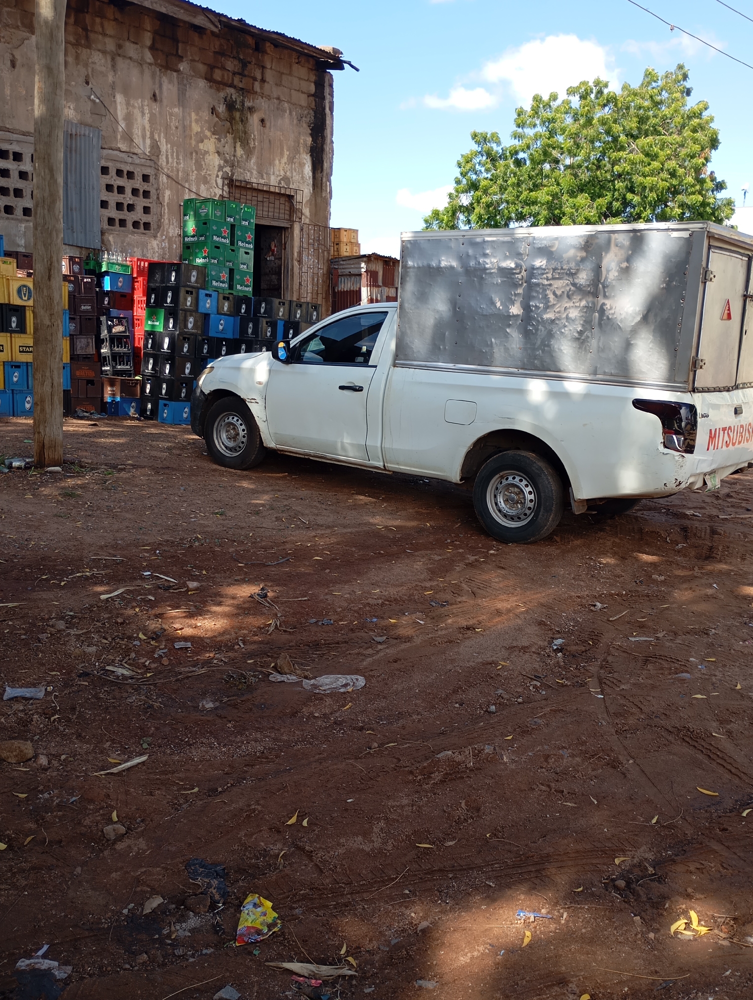 The image captures an outdoor scene in Shendam, Nigeria, likely a commercial or storage area. In the foreground, a white Mitsubishi pickup truck with a silver cargo box is parked on a dirt and gravel ground. The truck shows some wear and tear, with visible dents and scratches on its rear bumper and body. The cargo box appears to be made of corrugated metal.
Behind the truck, a large stack of colorful crates is piled high against a dilapidated building. The crates are mostly green, blue, and black, suggesting they hold beverages. Some of the green crates are identifiable as Heineken. The building behind the crates has a rough, textured exterior, with visible signs of aging and disrepair, including peeling paint and a corrugated metal roof. A doorway and a window with a perforated concrete block design are visible on the building's facade.
To the right of the building and in the background, a lush green tree with dense foliage stands tall against a bright blue sky dotted with white clouds. The ground is covered with reddish-brown soil, scattered with debris, including fallen leaves, small stones, and plastic waste. Tire tracks are visible in the dirt, indicating recent vehicle movement. The overall impression is one of a functional, perhaps somewhat unkempt, commercial area in a Nigerian city.