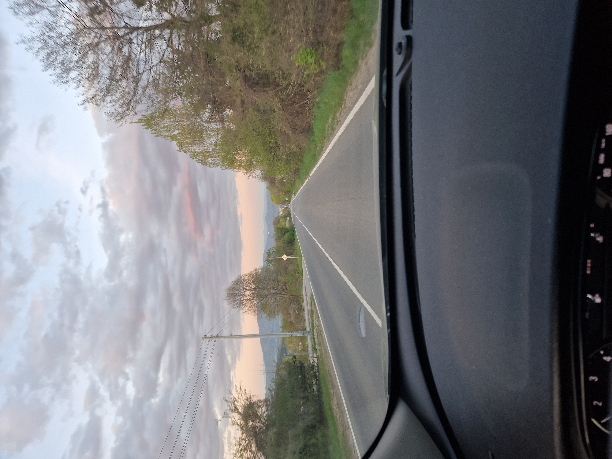 The image depicts a view from inside a vehicle traveling on an asphalt road. The road has a painted white line separating lanes. Green vegetation and trees line both sides of the road. Power lines and poles are visible adjacent to the road. In the distance, there are hints of mountains. The sky is filled with clouds, some tinted with pink hues. The interior of the vehicle is visible, including part of the dashboard and a gauge cluster. The location appears to be a rural or semi-rural area. City: Unknown, Unknown.