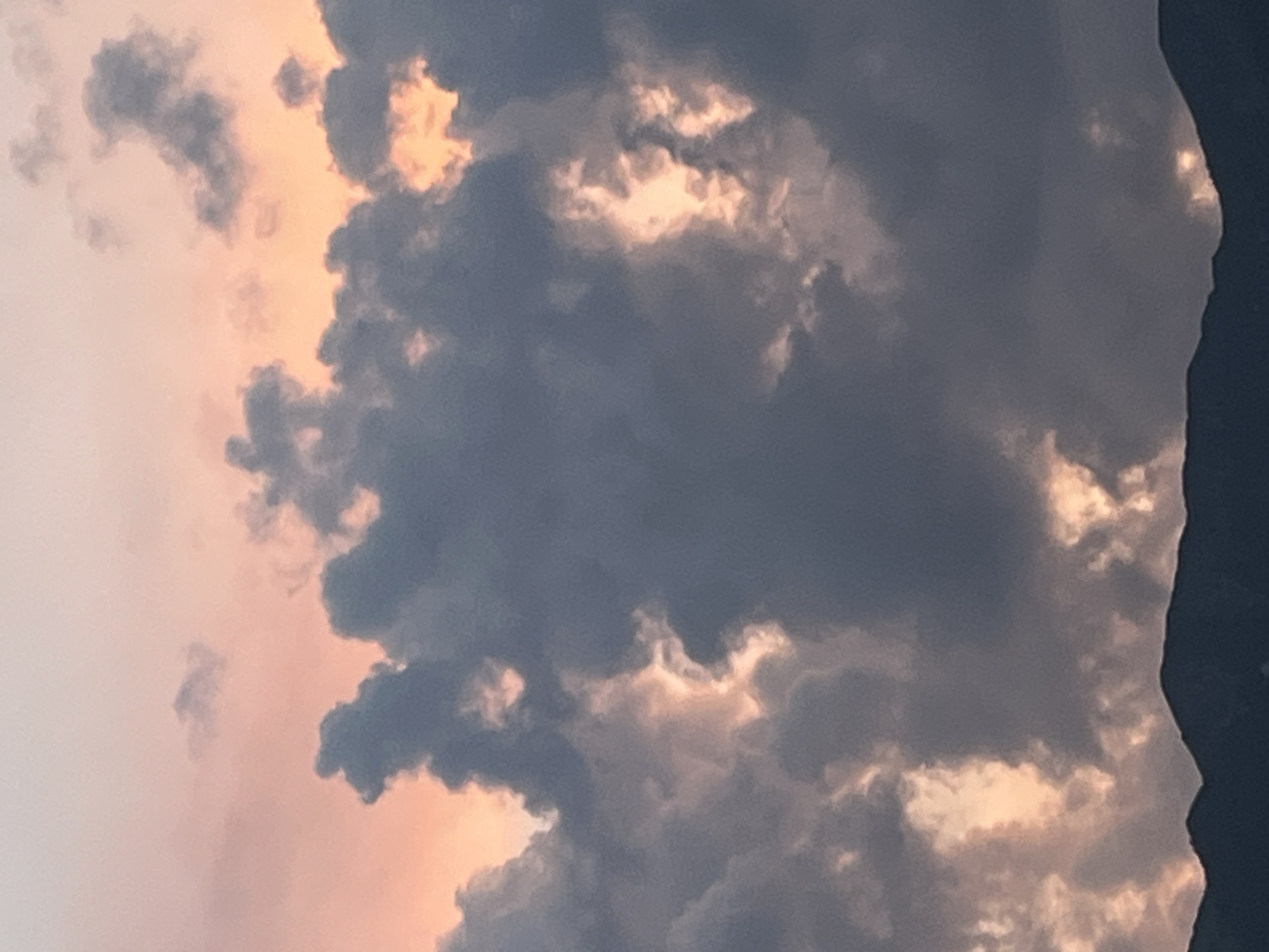 This image depicts a dramatic sky over the silhouette of mountains, likely the Alps, near Garmisch-Partenkirchen, Germany. The sky is a blend of deep grays and blues from thick clouds, with pockets of vibrant orange and pink light breaking through, suggesting either sunrise or sunset. The clouds themselves are textured and appear to be actively lit from behind by the unseen sun, creating areas of intense brightness and deep shadow. The mountain range at the bottom of the frame is rendered as a dark, undulating line against the colorful sky, its peaks softly defined. There are no people, animals, or man-made structures visible in the image, focusing solely on the natural elements of the sky and mountains.