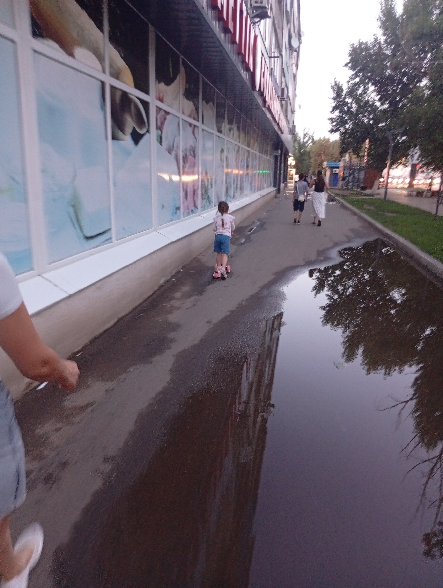 The media file depicts an outdoor scene on a sidewalk next to a building. The building has large, reflective windows displaying what appear to be advertisements or murals. The sidewalk is wet, with a large puddle reflecting the surrounding environment, suggesting recent rain or a moist day.
Several individuals are present in the scene. In the foreground, partially visible from the waist down, is someone wearing a white shirt and blue shorts. Further down the sidewalk, a young girl is riding a pink scooter. Behind her, a couple walks away from the camera; the person on the left is wearing a grey t-shirt and dark shorts, while the person on the right is wearing a white skirt and dark top.
The time of day appears to be late afternoon or early evening, as indicated by the soft lighting and the sky's color. The weather seems mild, as people are dressed in light clothing. The setting is urban, with a building on one side and trees and a street visible in the background. The overall mood is casual and everyday, capturing a moment of activity on a city street. There is no visible text in the media file.