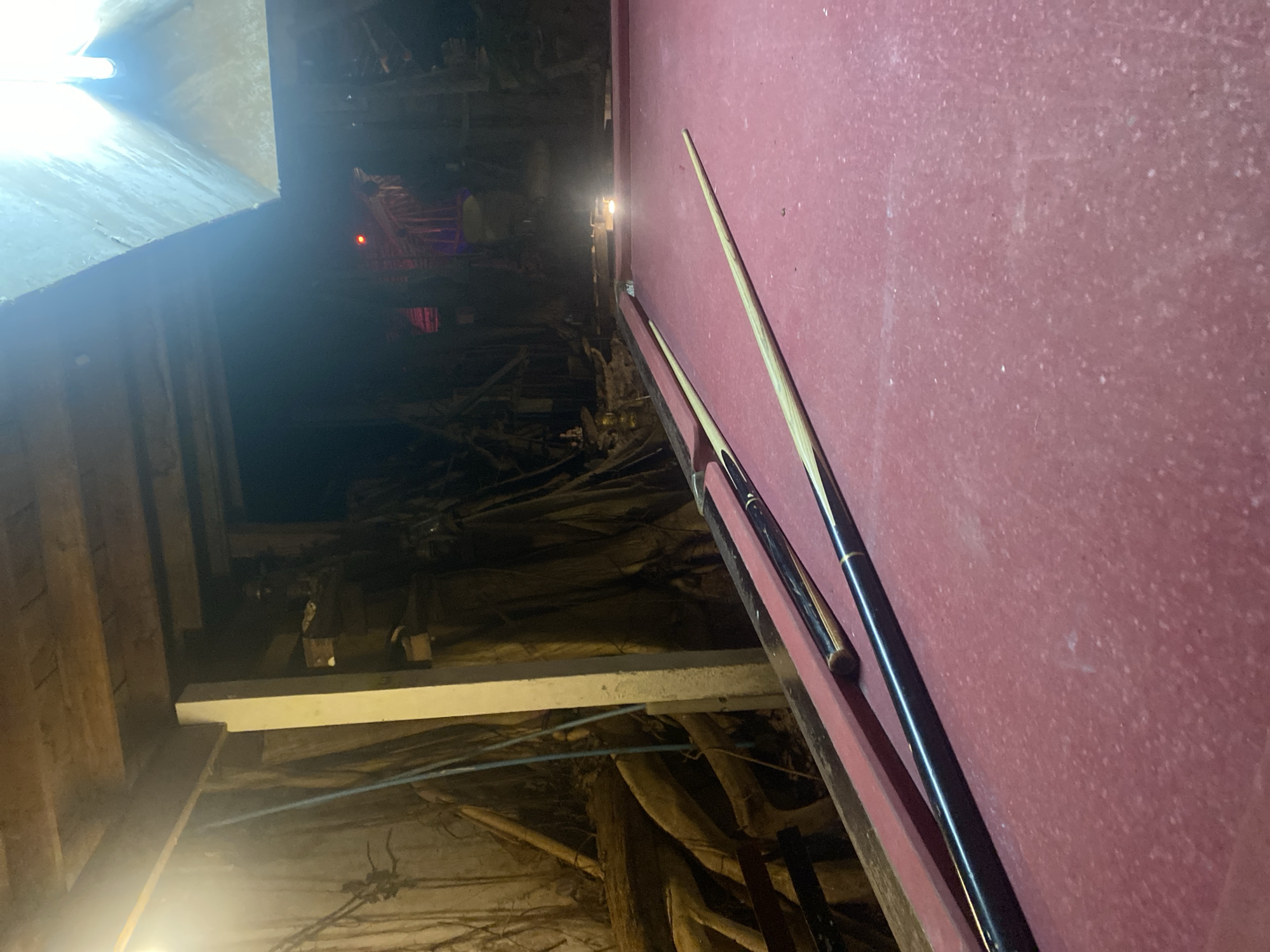 A tilted shot focuses on a red-felted pool table and two pool cues resting on its edge. To the left of the pool table, an obstructed and dark space reveals structural wood and beams, along with a glimpse of red lights in the distance. In the background, an indeterminate area appears cluttered and disorganized. The location is identified as Baan Don Sai, Thailand.