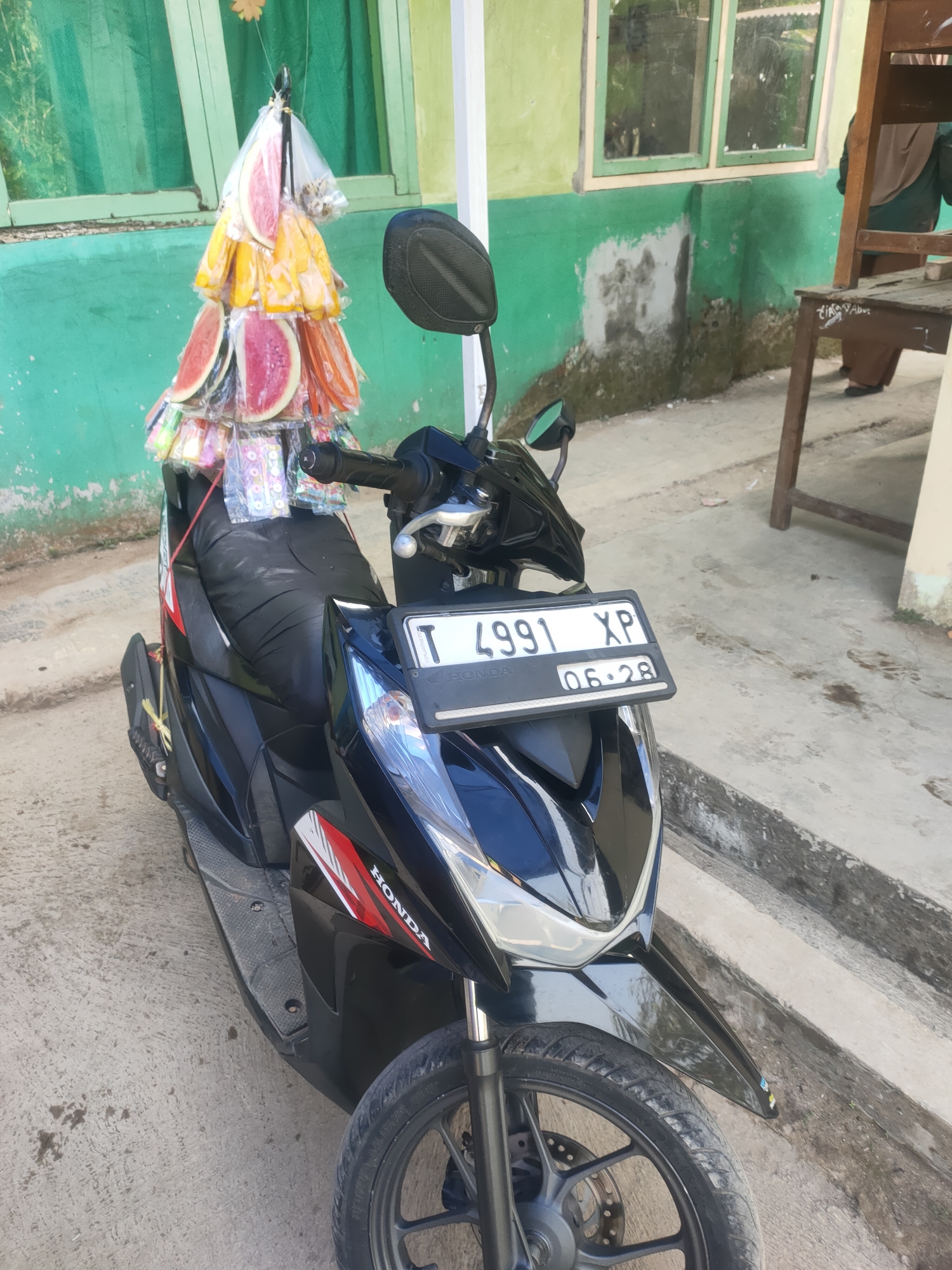 A black Honda scooter with a license plate reading "T 4991 XP 06.28" is parked on a concrete surface. Attached to the handlebars of the scooter is a decorative display of what appears to be sliced fruit, possibly watermelons, individually wrapped in plastic and arranged in a pyramid shape. The scooter is positioned in front of a building with a green wall and windows. In the background, a wooden chair is visible. The ground is made of concrete, and there are some dark stains present.