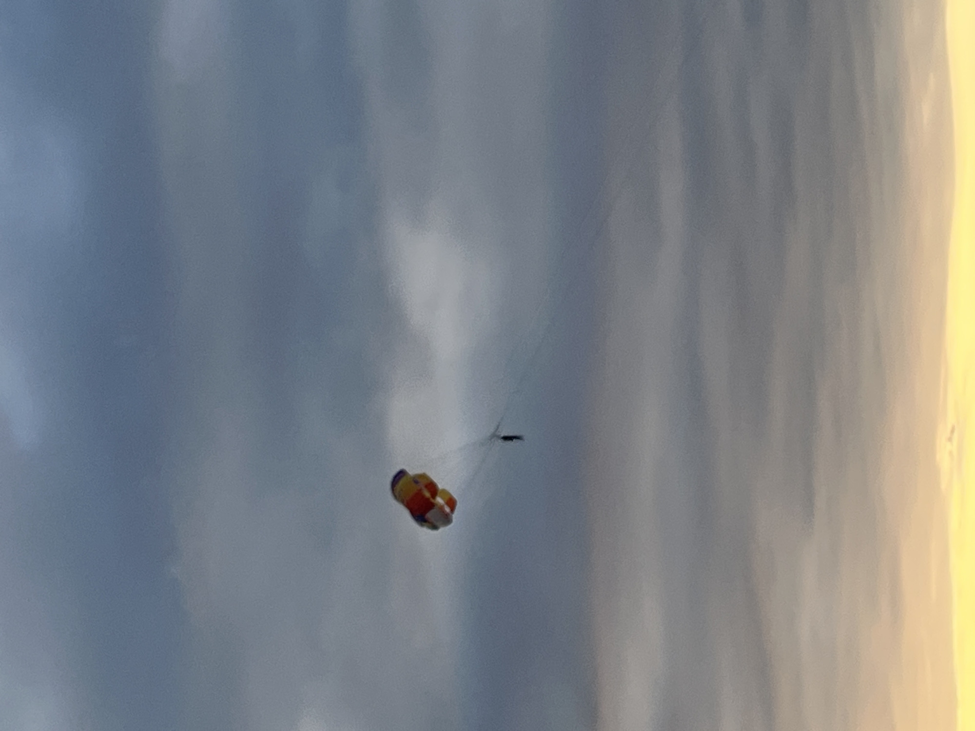 In Ureki, Georgia, a person is parasailing against a backdrop of a cloudy sky with a hint of yellow and orange at the horizon, suggesting either sunrise or sunset. The person is suspended in the air, attached by a rope to the colorful parachute. The parachute is predominantly yellow, red, and blue, and appears to be inflated and actively guiding the person's flight. The cloudy sky is a mix of dark gray and lighter blues, creating a dramatic contrast with the bright colors of the parachute.