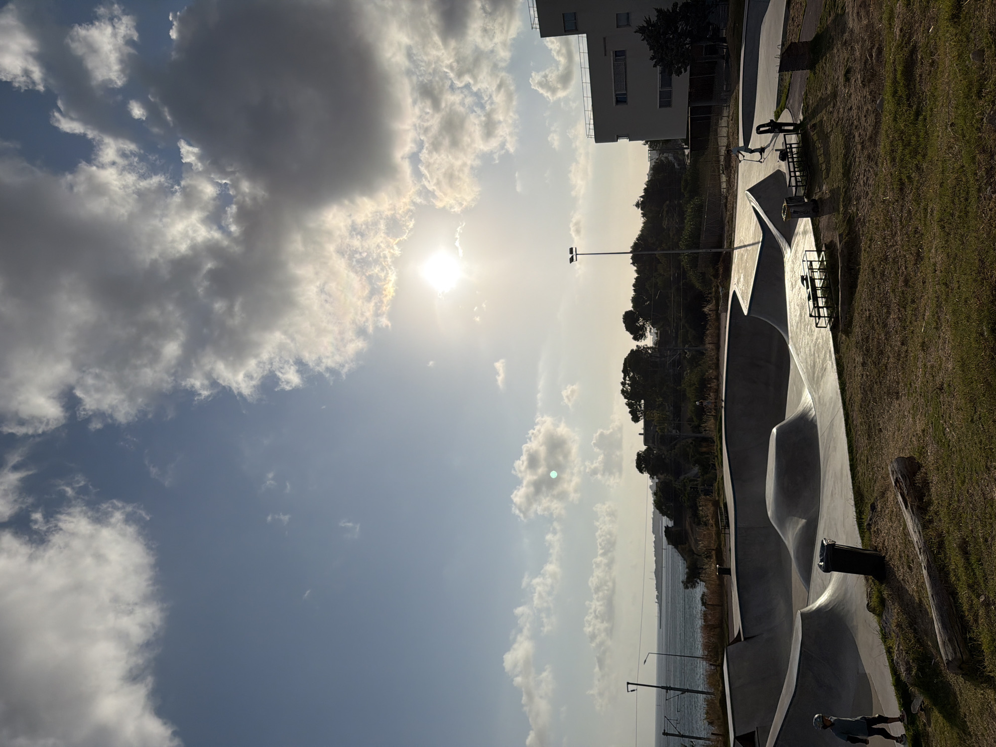 The image depicts a skatepark located by the sea, likely in Cascais, Portugal, given the distinctive architecture of a modern, white, multi-story building visible in the background to the right, which resembles buildings found in the area. The sun is shining brightly in the sky, partially obscured by clouds, creating dramatic lighting and casting long shadows.
In the foreground, a young person wearing a helmet and knee pads is standing near a skateboard on the grass. The skatepark itself is a concrete structure with various bowls, ramps, and obstacles. Further into the skatepark, another person is riding a scooter. Two trash receptacles are visible within the skatepark.
Beyond the skatepark, a body of water, presumably the Atlantic Ocean, stretches to the horizon. A line of trees and vegetation separates the skatepark from the water. A lamppost stands tall in the middle ground. In the distance, a low-rise building with a flat roof is visible. The overall scene suggests a public space designed for recreational activities, bathed in the warm light of either early morning or late afternoon.
