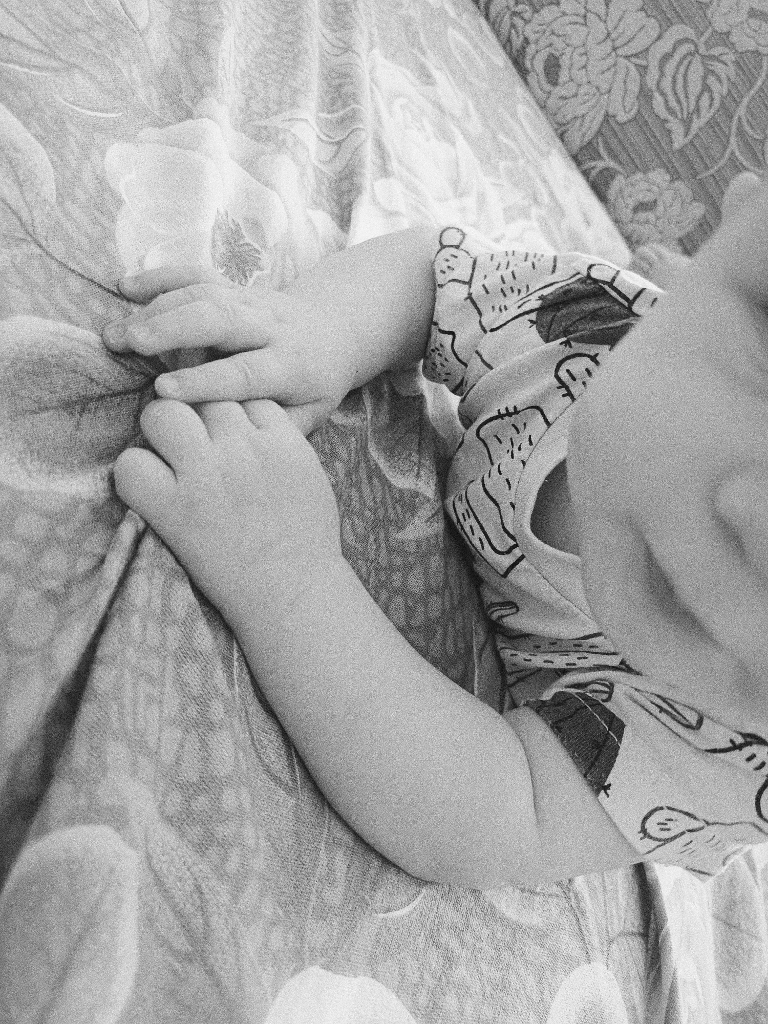 A black-and-white close-up photograph shows a baby's hands and arms resting on a patterned fabric surface. The baby is wearing a patterned short-sleeved shirt. The background features two distinct fabric patterns: one with floral motifs and another with a less discernible design. The absence of color and the close proximity suggest an intimate moment captured. The location, Tobolsk, Russia, is an external assumption, not directly discernible from the image itself.