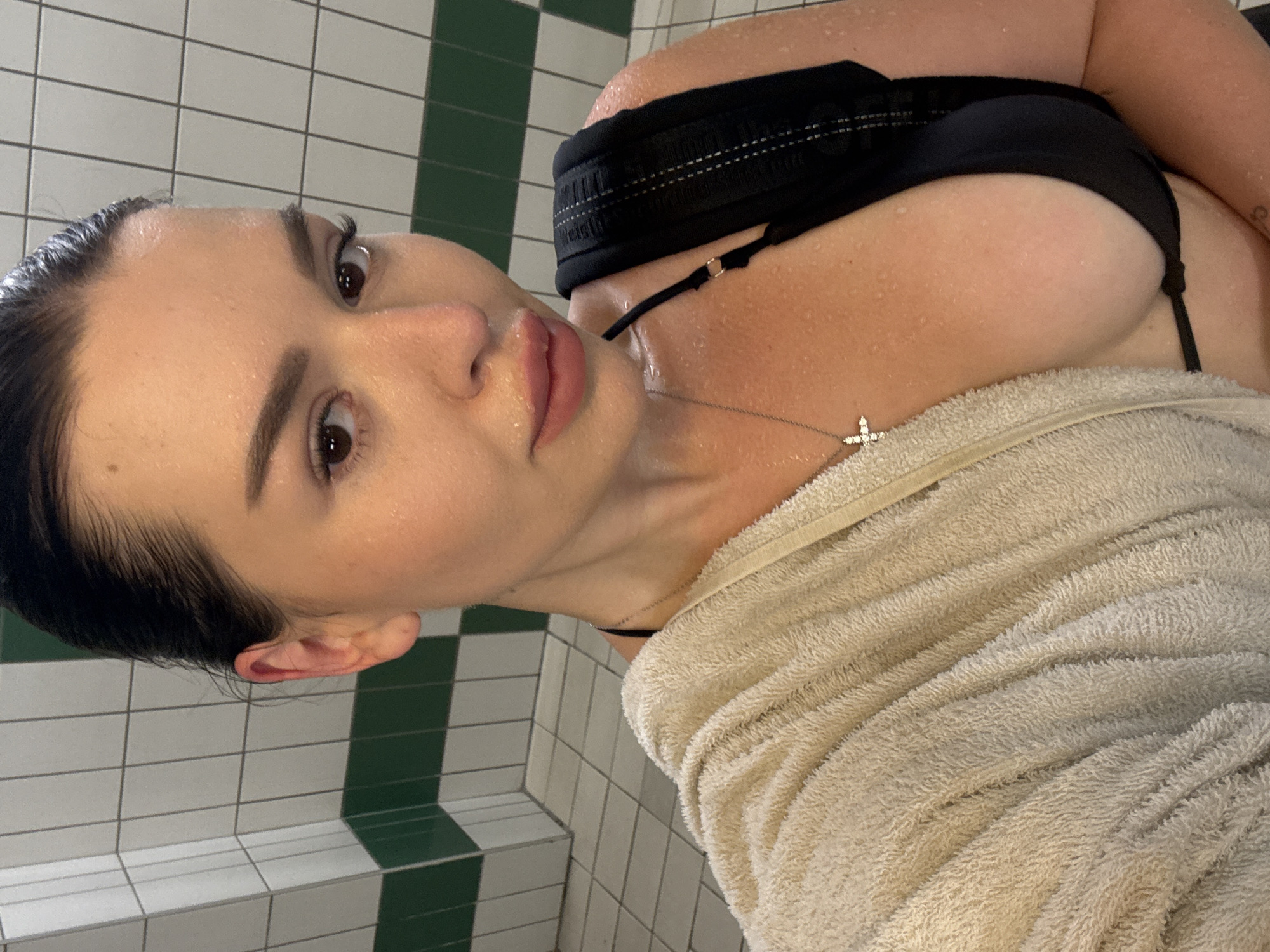 A woman, identified as a 33-year-old Czech female, is seen in a selfie taken in a locker room. She is wearing a black bikini top and has a cream-colored towel draped over her left shoulder and chest. A silver necklace with a cross pendant is visible around her neck. The background consists of tiled walls, with alternating white and green tiles, and a tiled floor. Water droplets are visible on her skin, suggesting she may have recently showered or been in a humid environment. The overall setting appears to be a public or communal shower area. The image does not provide any specific landmarks or details that would allow for pinpointing a location within Prague, Czechia, beyond it being a locker room.
