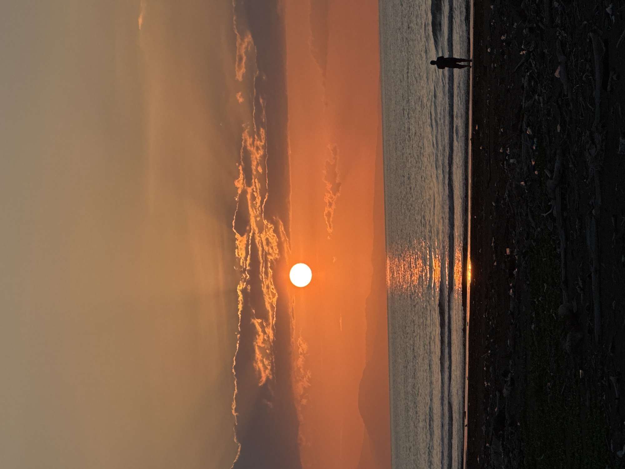 An image depicts a sunset over a body of water with a beach in the foreground. The sun, a bright orange orb, is positioned in the upper center of the frame, partially obscured by clouds that are illuminated by its light. Rays of sunlight emanate from behind the clouds. The sky transitions from a deep orange near the horizon to a lighter, more muted orange at the top of the image. In the distance, silhouetted mountains or hills are visible against the sky. The water is calm, with gentle waves rolling towards the shore, and a shimmering reflection of the sun stretches across its surface. On the beach, which is dark and covered with debris, a lone figure stands facing the water. The figure is a silhouette, appearing to be an adult. The overall scene is one of natural beauty and tranquility, with the setting sun casting a warm glow over the landscape. This scene is located in the city of Meunasah Jurong, Indonesia.