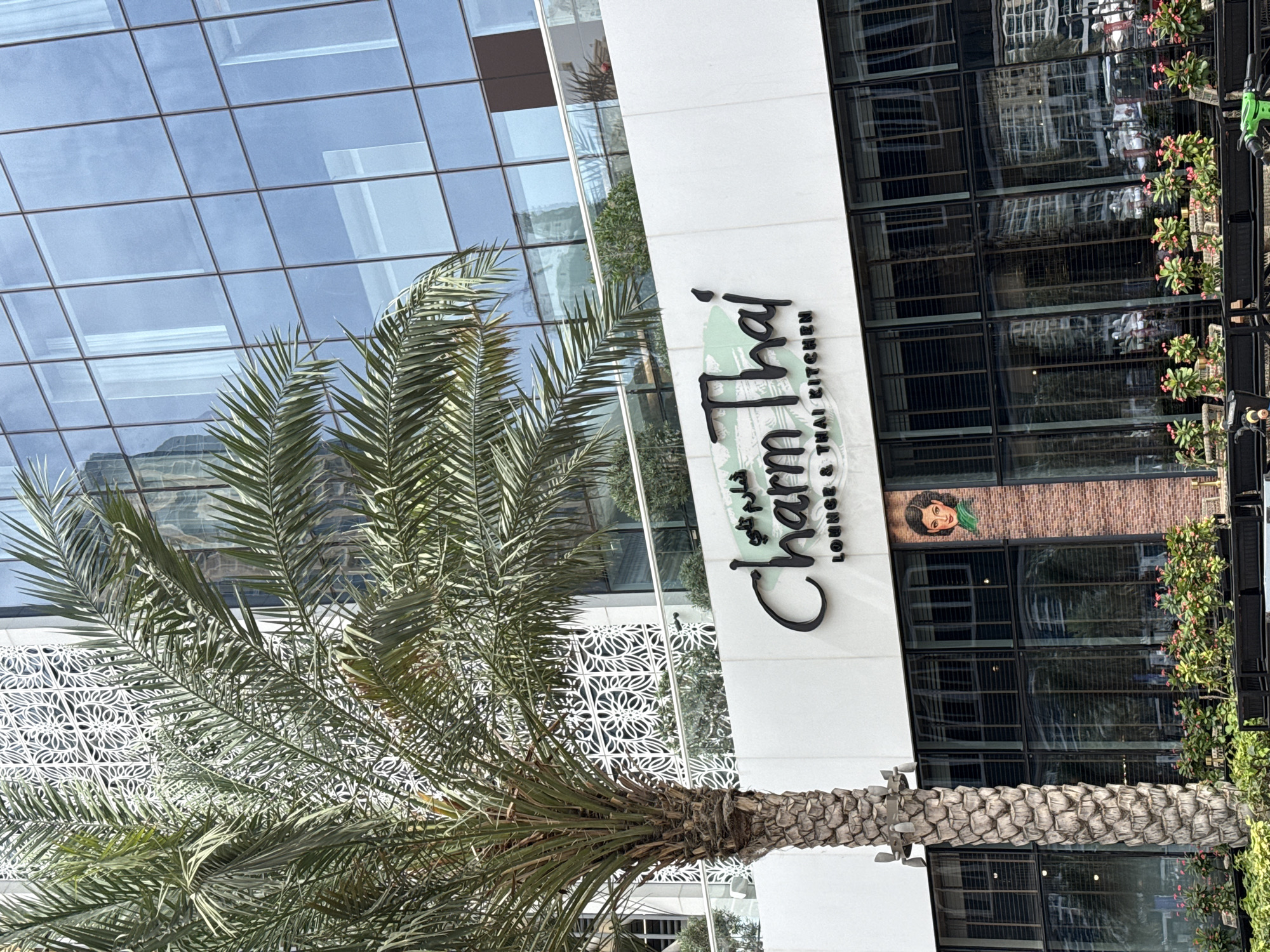 The image shows the exterior of a building in Dubai, United Arab Emirates. A tall, multi-story building with a glass facade is partially visible on the left side of the frame. Adjacent to the glass building is a white structure with the signage "Charm Thai Lounge & Thai Kitchen" prominently displayed. A mature date palm tree is situated in front of the building, partially obscuring the view. The right side of the image shows part of the restaurant's exterior, including windows and plants. Above the windows, there is a decorative element featuring an image of a woman with a green headscarf. The restaurant seems to have an outdoor seating area with chairs and tables.