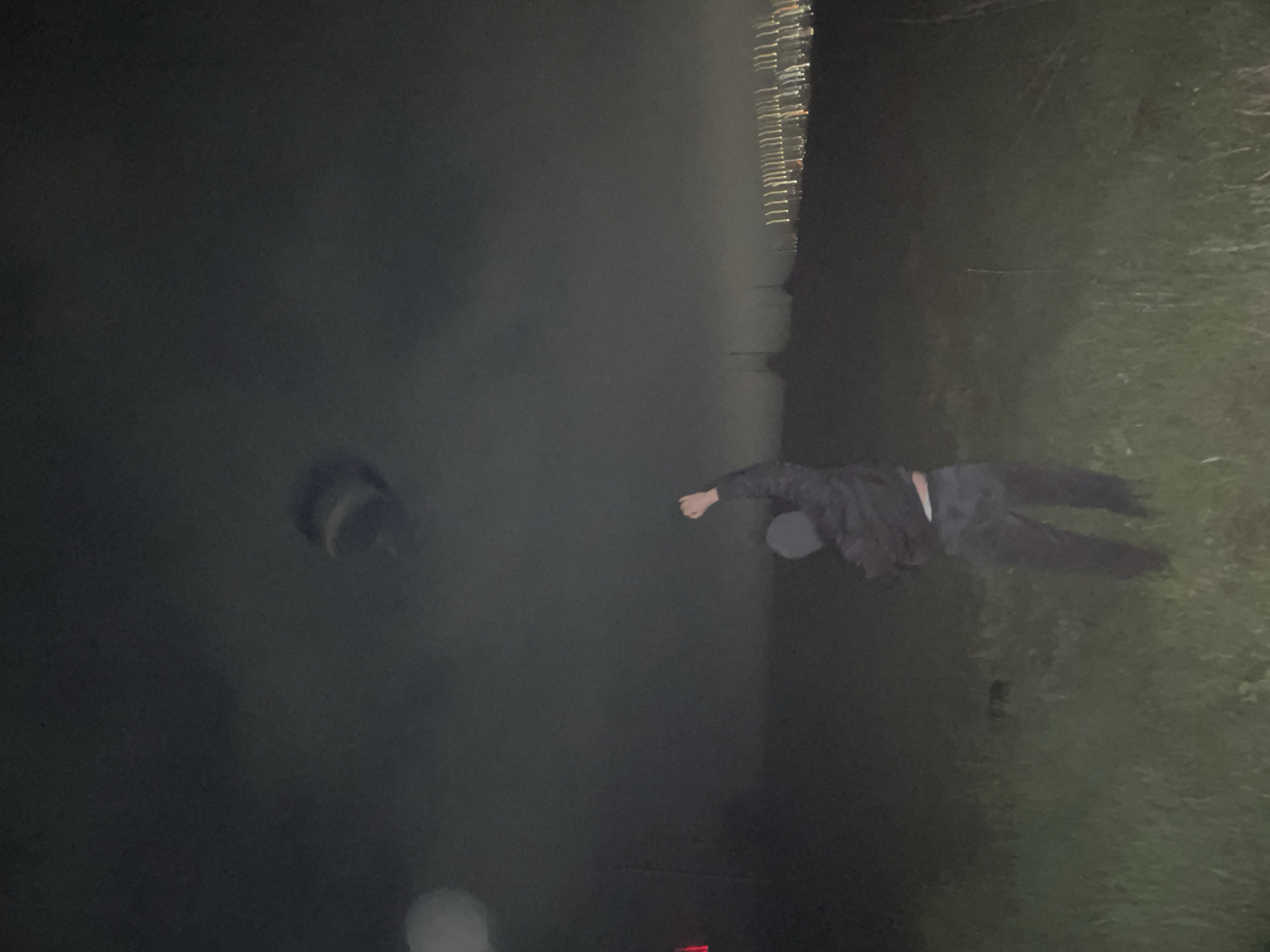 A person lies prone on grass at night. A dark body of water or fog is on the left side of the image. In the distance, blurred lights possibly represent a city skyline. The image's location is stated to be Razgrad, Bulgaria, but the information is not visually confirmed.