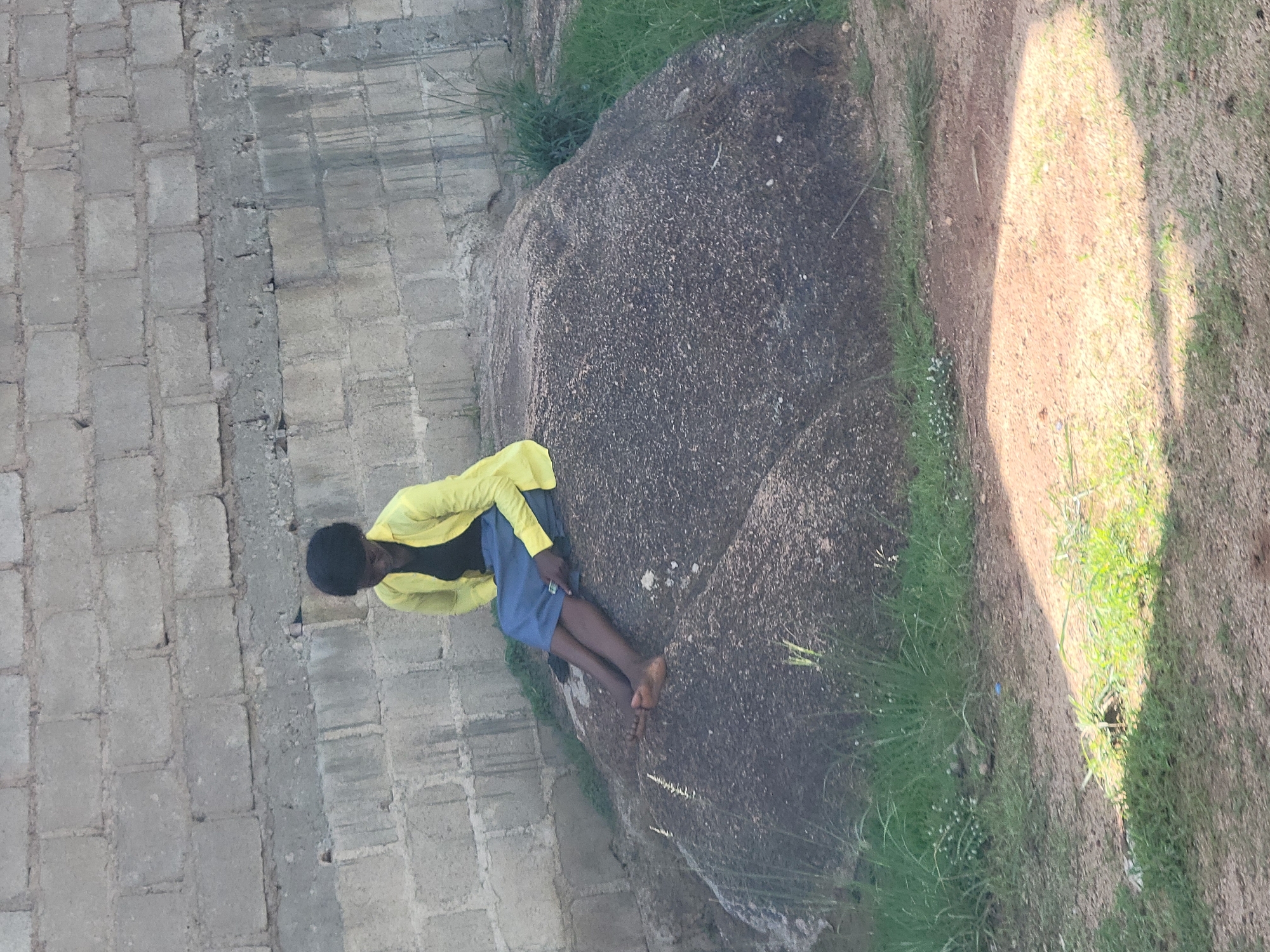 A young Black female wearing a yellow blouse and a blue skirt is sitting on a large, rough-textured rock. Her legs are crossed, and her feet are bare. She is looking down, holding a dark object, possibly a phone, in her right hand. The rock is situated outdoors, with grass and dirt visible in the foreground. In the background, a wall constructed from grey cinder blocks is visible. The lighting suggests it is daytime, with sunlight casting shadows on the ground. The overall setting appears to be an unpaved area, potentially on the outskirts of a city, given the combination of natural elements and rudimentary construction. It is not possible to determine the specific location within Jos, Nigeria.