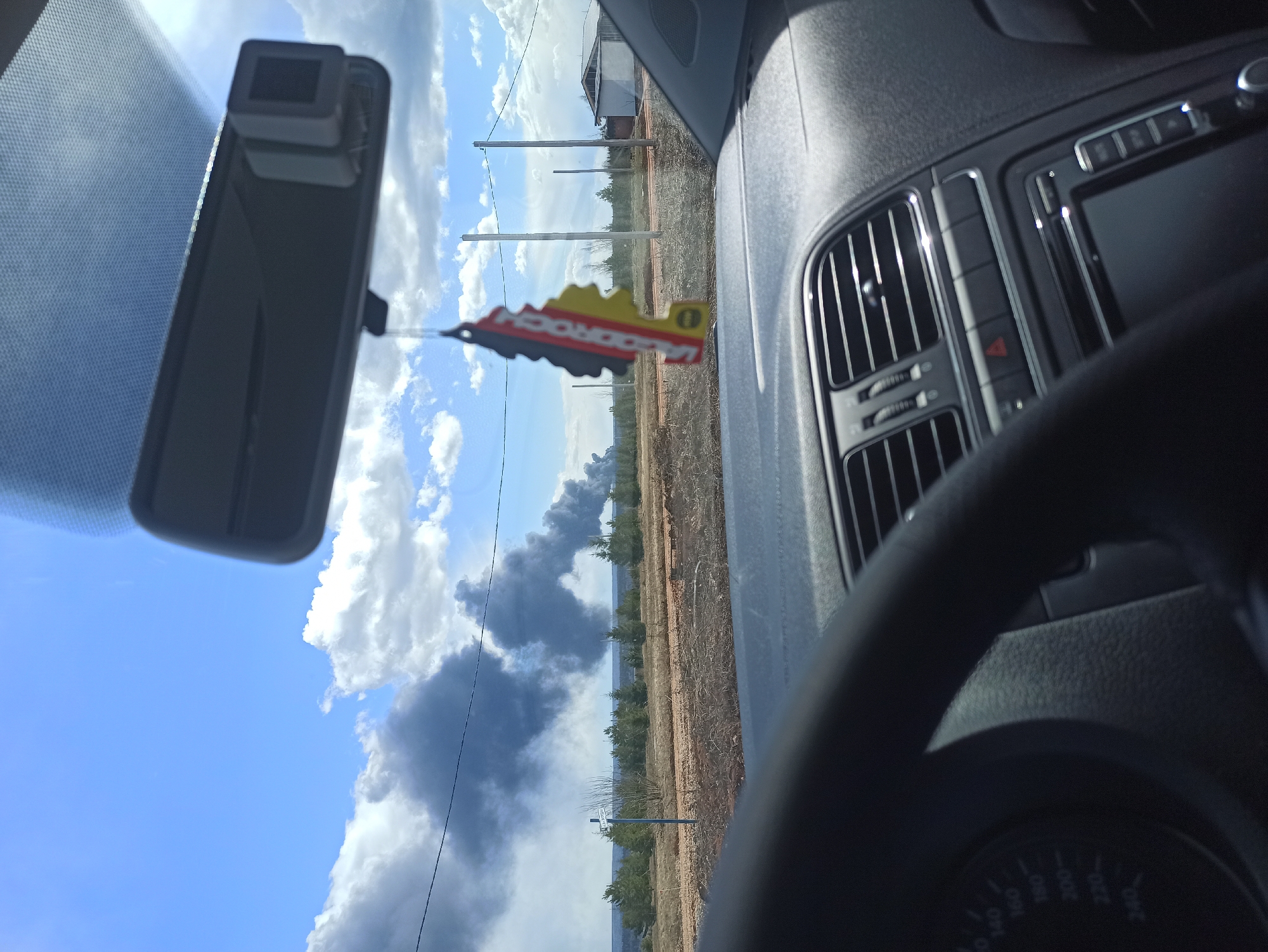 The image, taken from inside a car and rotated 90 degrees counter-clockwise, depicts a view through the windshield towards a rural landscape under a partly cloudy sky.
In the foreground, elements of the car's interior are visible. On the right, the black dashboard features air conditioning vents with chrome accents, several buttons (including a prominent red hazard light button), and part of an infotainment system screen. A segment of a black steering wheel is visible at the bottom right, partially obscuring a speedometer which faintly shows numbers such as "160," "180," "200," "220," and "240" (likely km/h). In the upper left, a rearview mirror is prominent, with a black rectangular device, possibly a dashcam, mounted on top. Hanging from the rearview mirror is a key-shaped air freshener, predominantly red and yellow, with the text "JADOROCK" discernible.
Beyond the car, the exterior scene unfolds under a bright blue sky dotted with white cumulus clouds and darker grey clouds. The landscape consists of a wide, reddish-brown dirt road or field stretching into the distance, bordered by several wooden utility poles. A line of green trees, possibly pines, forms the horizon, and a small, white building with a dark roof is visible in the far distance on the right.
A significant event is occurring in the distance: a massive, dark grey plume of smoke is rising from behind the treeline, indicating a large fire. The smoke extends high into the sky, partially obscuring the clouds. The scene suggests a daytime setting, likely in an open, undeveloped area.