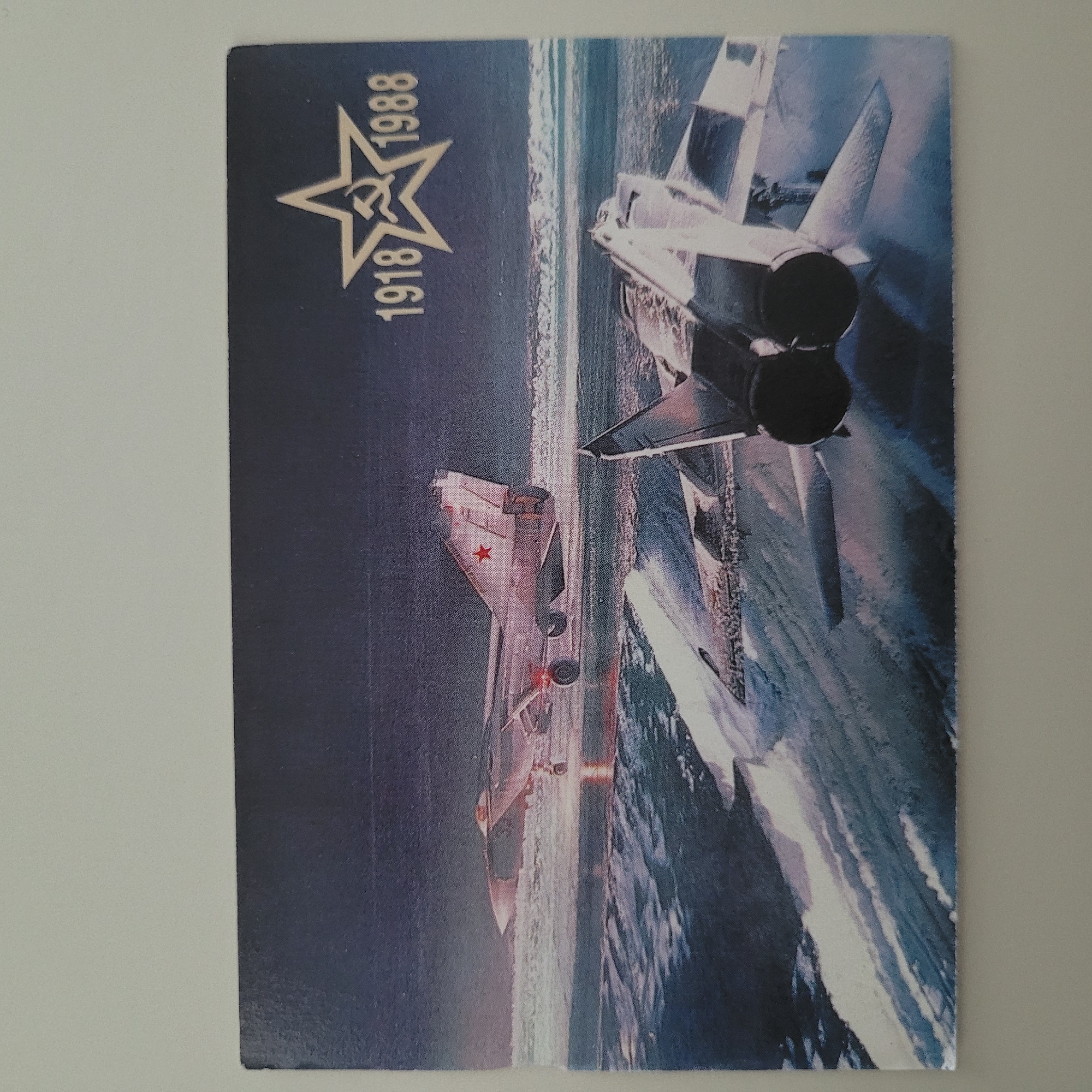 The image is a postcard featuring a depiction of two Soviet-era fighter jets. In the background, a jet with a red star on its tail is on a runway, appearing to be taking off or landing. In the foreground, the rear of another fighter jet is visible, showing its twin engines. Above the jets, on the right side of the postcard, is a prominent outline of a five-pointed star containing the hammer and sickle emblem, flanked by the numbers "1918" and "1988." The overall scene appears to be a night or dusk setting over what looks like a water or icy surface, with spray or mist rising. There are no people visible in the image. The postcard itself is the subject, not a scene from Jūrmala, Latvia.