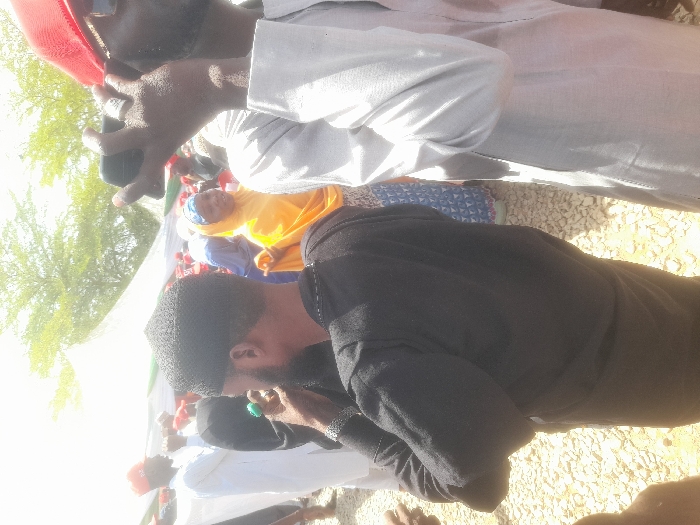 The image shows a group of people gathered outdoors in Gusau, Nigeria, at coordinates 12.156, 6.709. The scene is primarily focused on three men, all wearing traditional clothing. One man, wearing a black top, is holding something in his hand, possibly a green bottle, and looking to his left. Another man, standing directly behind him, is wearing a grey shirt and a red cap. He is holding a black smartphone in his right hand and looking toward the left side of the frame. The third man, wearing a white top, is standing further back. The crowd of people is partially obscured by a large white tent. The environment is sunny and the ground is covered in loose gravel.