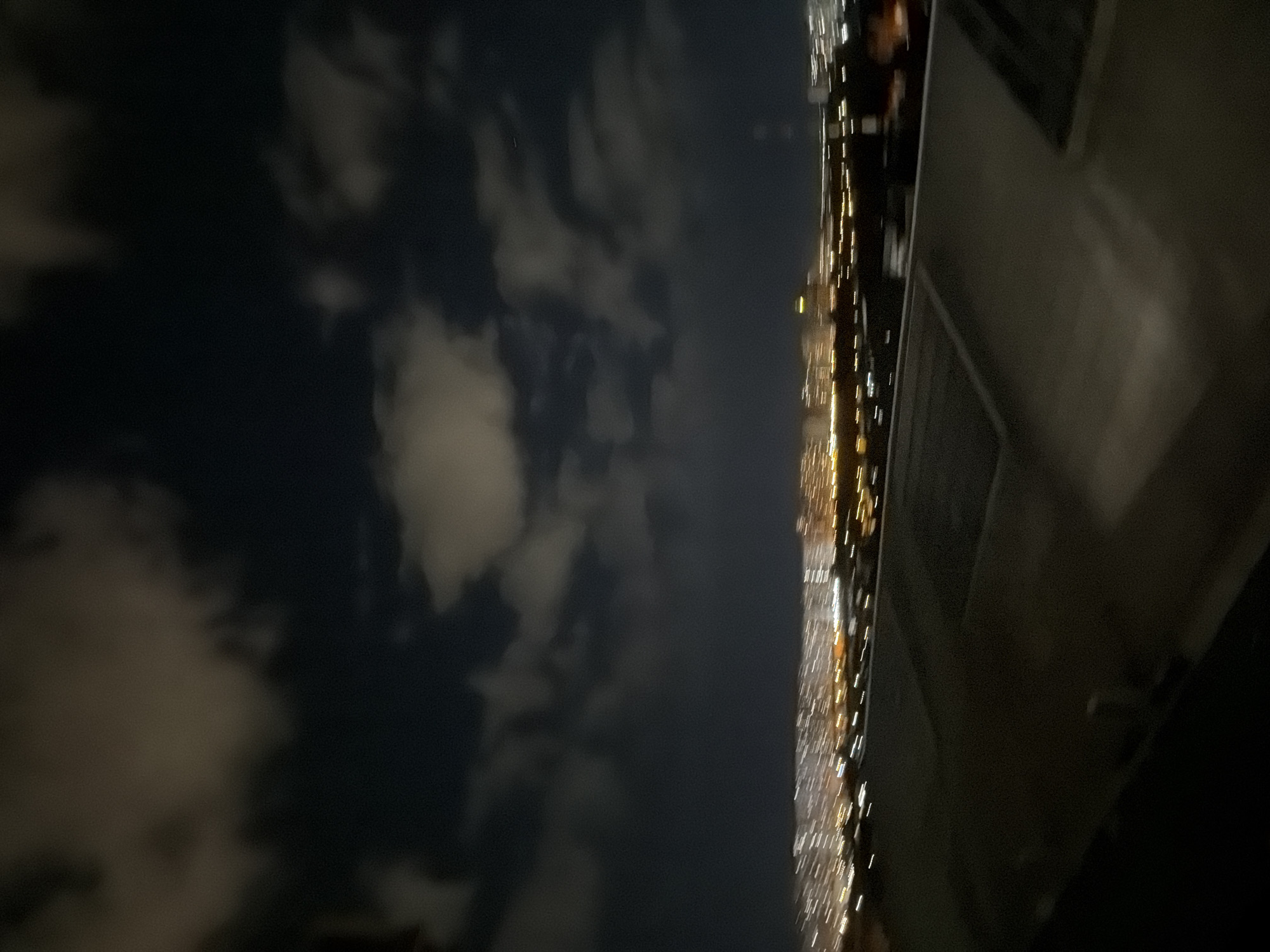 The image depicts a blurry night view of a cityscape with clouds in the sky. In the foreground, there is a dark, textured surface that appears to be a rooftop. Beyond the rooftop, a city skyline is visible, illuminated by numerous lights that are stretched horizontally due to motion blur, indicating a long exposure or camera movement. The lights suggest a densely populated urban area with buildings and possibly roads. The sky is dark, with patches of lighter clouds that are also blurred. The overall impression is one of a nocturnal urban landscape viewed from an elevated position, likely a rooftop. The location is specified as Kyiv, Ukraine. There are no discernible people, specific objects, or actions taking place.