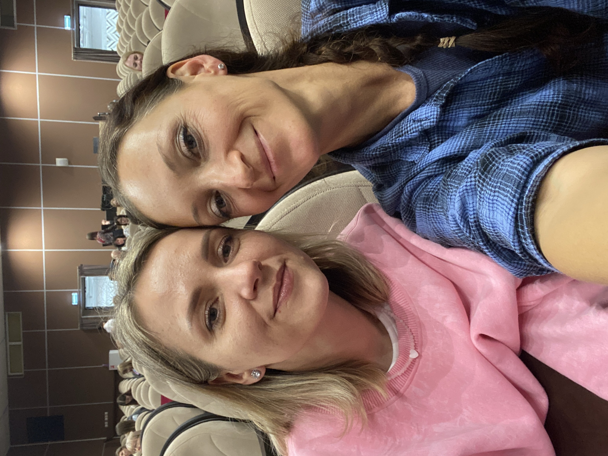 Two women pose for a selfie indoors. The woman on top has brown hair pulled back, and wears a blue plaid shirt. The woman on the bottom has blonde hair, and wears a pink sweatshirt. They are seated in an auditorium with rows of upholstered seats. Other people are seated in the background. The location is Ulyanovsk, Russia.