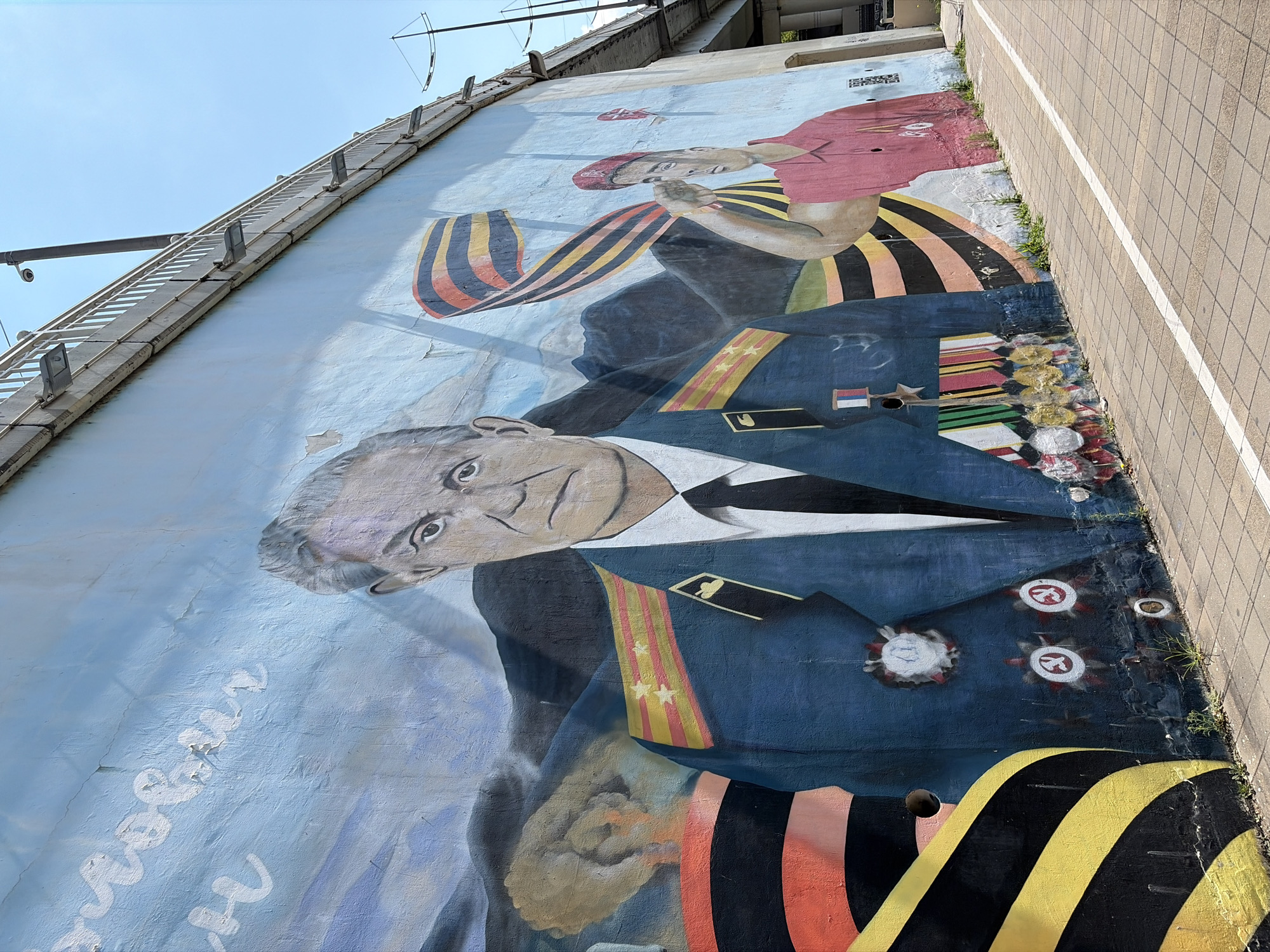 The image depicts a mural painted on a concrete wall in Sochi, Russia. The mural features a central figure of a man in a military uniform, adorned with medals and epaulets. To the right, another figure, a young man in a red shirt and beret, salutes. Behind them, there are elements suggesting a mountainous landscape and a Russian flag. The St. George's ribbon, a symbol of military valor, is prominently displayed in the mural. The wall is located alongside a pedestrian walkway with a white line marking a path. Overhead, a bridge structure with railings and lighting is visible against a clear blue sky. The mural appears to be a patriotic commemoration, likely related to a significant historical event or military figure.