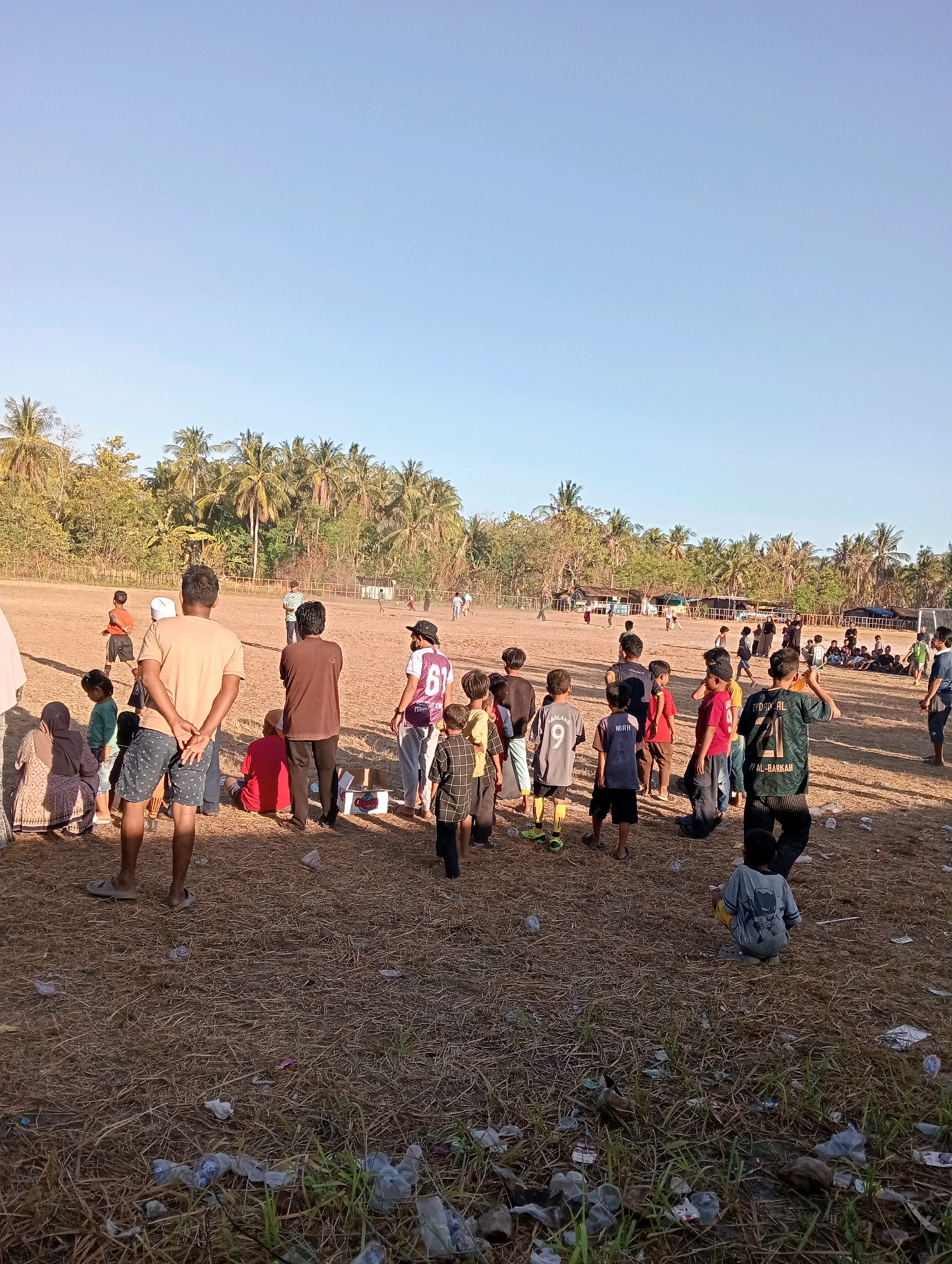 The image depicts an outdoor gathering of people, primarily children and adults, on a dry, dusty field surrounded by palm trees and vegetation, characteristic of Pagerungan Kecil, Indonesia. A group of young boys are engaged in a football match in the background, while others observe the game or interact in smaller groups. Several adults are standing and watching, some with their hands behind their backs. The ground is covered in dry grass and scattered with litter, including plastic bottles and wrappers. The sky is a clear, pale blue, indicating daylight. The overall scene suggests a community event or casual sporting activity in a rural Indonesian setting.