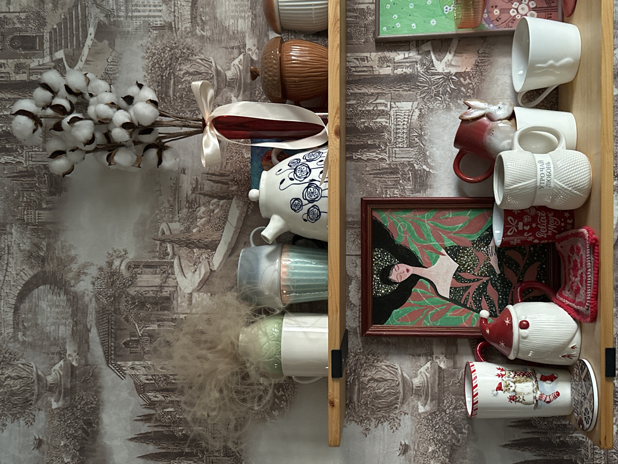 The image depicts a shelf in an interior setting, likely a home, adorned with decorative items and mugs. The background features wallpaper with a sepia-toned landscape of classical architecture and gardens, evoking an old-world aesthetic.
On the top shelf, several objects are arranged: a bunch of dried cotton plant stems in a red vase tied with a pale ribbon, a white teapot with blue floral patterns, a brown acorn-shaped ceramic container, and a ribbed white ceramic container. To the left, there are three stacked mugs, two of which are light blue and pink, and a white mug holding a fluffy, light-colored organic material.
The lower shelf displays an assortment of mugs and a framed artwork. The artwork is a portrait of a woman with dark hair and green foliage, in a dark, richly patterned dress. To the left of the artwork are two festive mugs: one is a tall, white cylindrical mug with a Christmas-themed design of a cat in socks and a candy cane border, and another is a white mug shaped like a Santa Claus gnome. A small round coaster with a mosaic pattern is placed in front of the Christmas mugs. In the center and right of the lower shelf are several more mugs: a red mug with a white rabbit figurine, a white mug designed to resemble a ball of yarn with Russian text that translates to "Morning Tea Love", a plain white mug, and a white mug with a subtle ribbed pattern. A small crocheted pink and red mat is placed near the Santa mug.
The lighting suggests an indoor environment, possibly with natural light coming from a window, casting soft shadows on the shelf. There are no people visible in the image, and no specific actions are depicted beyond the arrangement of objects. The overall impression is one of a curated display of decorative and functional items.
This image does not contain any specific landmarks or contextual information that would allow for its identification or location within the city of Ulyanovsk, Russia. The background wallpaper depicts a generic classical landscape, and the objects themselves are not indicative of any particular geographical origin.