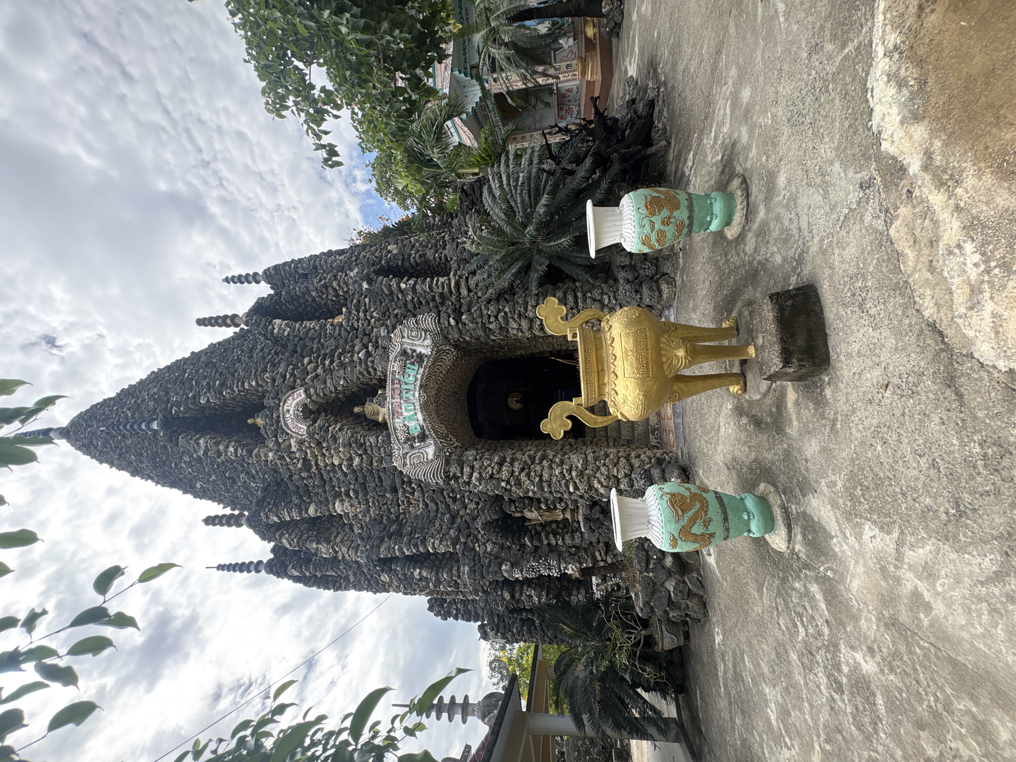 The image captures a unique, intricately constructed grotto-like temple or shrine made predominantly from dark, irregular stones, pebbles, or shells, set in Cam Ranh, Vietnam. The structure rises to a conical or tiered peak, adorned with multiple spire-like elements crafted from stacked circular components. An arched entrance, possibly the main doorway, features lighter-colored shell mosaics, with partially visible stylized text that appears to spell out "THICH CA" or similar, suggesting a Buddhist context.
In the foreground, two ornate decorative urns are placed on a concrete ground. To the left, a large, golden urn stands on stylized legs with an elaborate handle. To its right and further back, another urn is visible, light green and white with gold dragon motifs, standing on a pedestal. Lush green tropical foliage, including palm trees and leafy branches, frames the structure and rises behind it, hinting at a natural, possibly garden setting. The sky above is partly cloudy, indicating daytime, likely under bright, diffused light. No specific activity or people are depicted; the scene is static, showcasing the unique architecture and its surrounding environment.