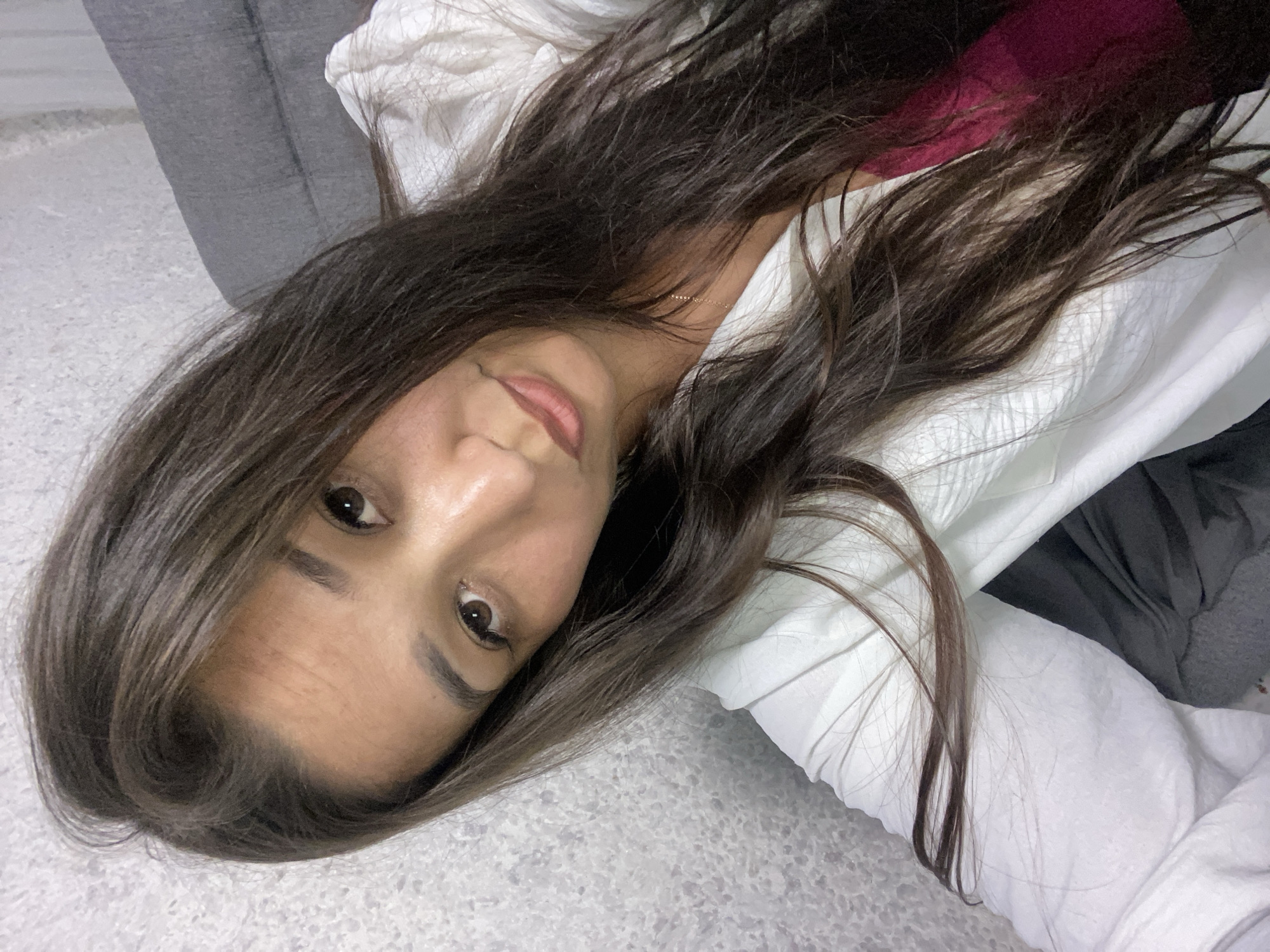 A woman with long, dark brown hair is shown in a selfie-style shot. She is wearing a white open jacket over a dark pink top. Her head is tilted, and her gaze is directed towards the camera. The background appears to be a textured grey wall and a portion of a grey sofa. The lighting is soft, and the overall scene suggests an indoor setting. There are no discernible landmarks or elements that would allow for specific identification of her location within the city of Ufa, Russia.