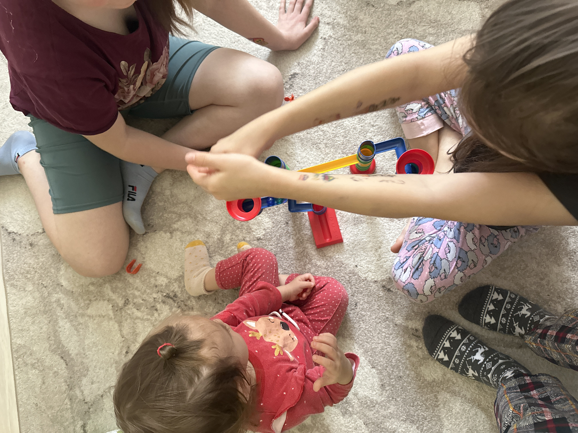 An overhead view captures three individuals, likely children, engaged in playtime on a light-colored, textured carpet indoors.
To the upper left, one person wears a maroon t-shirt, blue-green shorts, and light blue socks with the "FILA" logo visible on the left foot. Their arm, adorned with a temporary tattoo, extends towards the center. Opposite them, to the right, another individual, seen from above, wears pink pajamas patterned with white and blue sheep, and dark grey socks featuring a white Christmas tree and geometric design. Their arms, covered in several colorful temporary tattoos, are also directed towards the central toy.
At the bottom left, the youngest child lies or sits on the carpet, dressed in a red long-sleeved outfit with white polka dots and a deer design on the front. Their light brown hair is tied back in a small bun with a pink hair tie, and they wear cream-colored socks with yellow toes and heels.
A colorful children's toy, composed of red, blue, and yellow plastic components, likely a balancing toy or simple marble run, is central to the image, with the two older individuals appearing to be actively interacting with it. A small red U-shaped object lies on the carpet near the youngest child. The scene conveys a relaxed and focused atmosphere of shared play within a casual home setting.