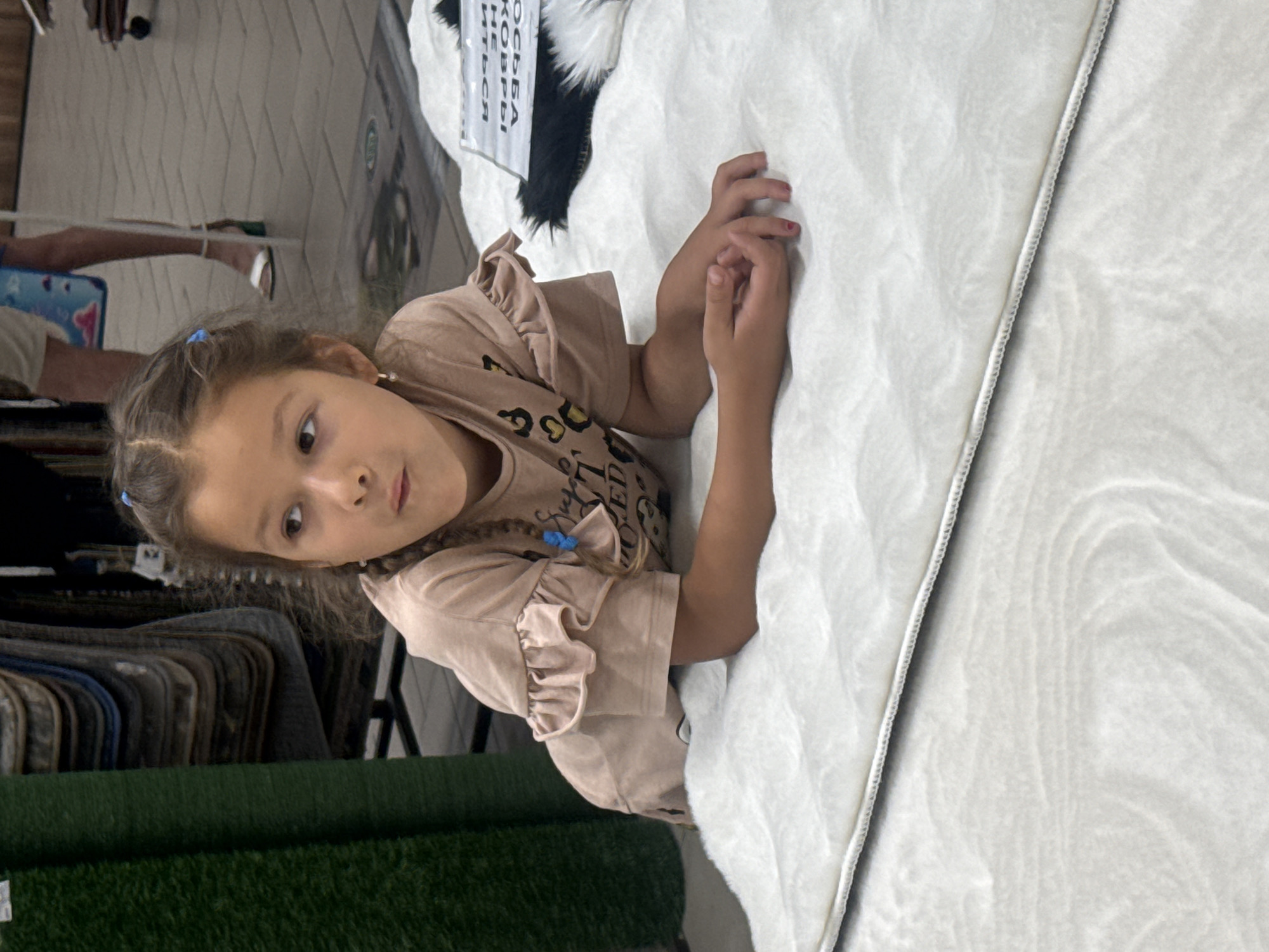 A young girl with braided brown hair and blue hair ties lies on a white rug in a store. She is wearing a light pink t-shirt with ruffled sleeves and a leopard print design on the front. Her arms are resting on the rug, and her hands are clasped together. In the background, there are rolls of green carpet and displays of other rugs. A sign with Russian text is visible on a black and white rug to the right of the girl. The floor appears to be tiled. The setting is consistent with a retail environment selling flooring or home goods.