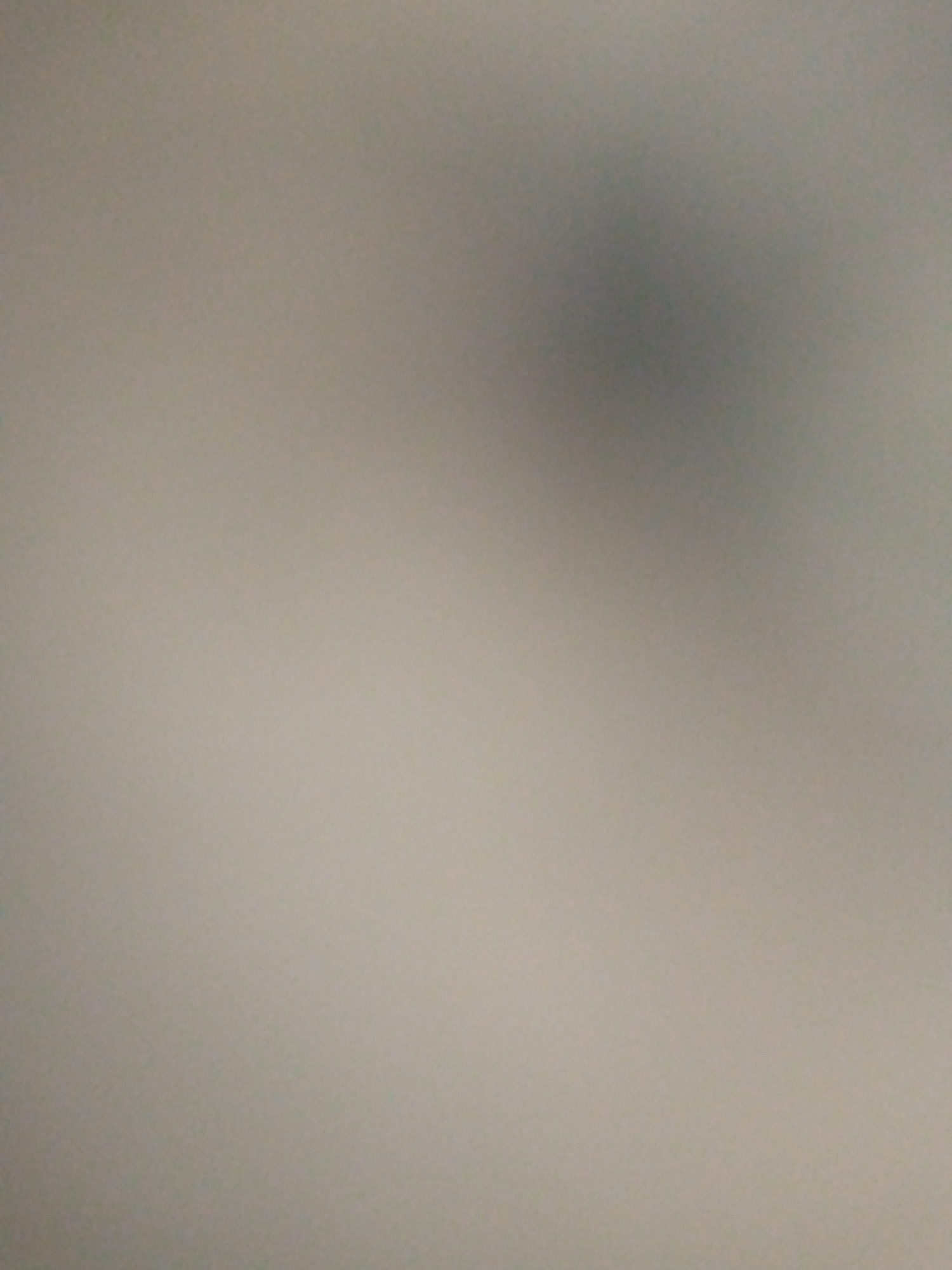 The image is a blurry, close-up shot of a neutral-colored surface, possibly a wall or fabric. There is a darker, undefined shadow in the upper right quadrant of the image. No specific objects, people, or discernible features are visible due to the lack of focus. The lighting appears to be diffused, creating a soft, even illumination across the scene. No location in Jalingo, Nigeria, can be determined from this image.