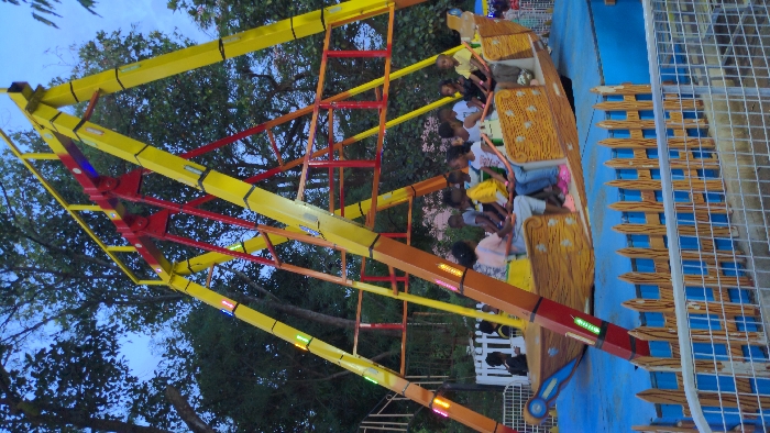 The image depicts an amusement park ride, identified as a "pirate ship" type, situated in the city of Moshi, Tanzania. The scene is captured during twilight or early evening, evidenced by the deep blue sky and the illumination of lights on the ride structure.
The central subject is a large, brightly colored pendulum ride. Its support structure consists of two prominent yellow A-frames connected by a red horizontal crossbar and pivot point. Numerous rectangular LED lights, exhibiting white, green, and yellow hues, are integrated along the yellow support beams and the orange swing arms of the ride. A brown, wood-textured boat-shaped gondola, with elevated bow and stern, is suspended from the red pivot.
Inside the gondola, approximately 8-10 children of varying ages (estimated between 5-12 years old) are seated in rows, facing inwards towards the center of the boat. They are wearing casual clothing. The gondola is observed in a slightly elevated position.
The ride is positioned over a blue ground surface, possibly designed to simulate water, which is enclosed by a decorative, light brown wooden-style picket fence. In the immediate foreground, a grey metal mesh fence is visible, separating the viewer from the ride area. The background features dense green foliage from trees, silhouetted against the dark sky. Faint figures are discernible near a white picket fence in the distant left background.