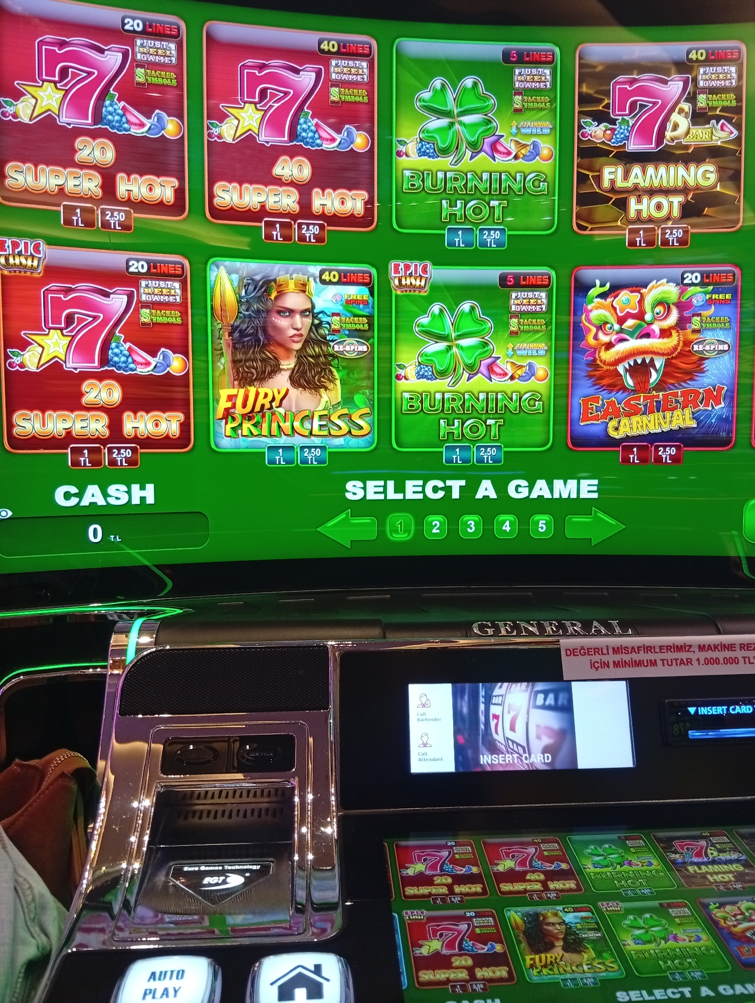 The image displays a close-up view of a slot machine screen in a casino, likely in Davlos, Northern Cyprus/Cyprus, given the on-screen text. The screen showcases various slot game titles with vibrant graphics, including "20 SUPER HOT," "40 SUPER HOT," "BURNING HOT," "FLAMING HOT," "FURY PRINCESS," and "EASTERN CARNIVAL." Below the game selections, a section labeled "CASH" displays "0 TL," indicating no current credit. Further down, "SELECT A GAME" is prominently displayed with numbered options and navigation arrows. A smaller screen on the lower right shows a prompt to "INSERT CARD" and options to "Call Bartender" and "Call Attendant." The surrounding area of the slot machine is metallic and appears to be well-maintained. The lighting is bright, typical of an indoor entertainment venue. No people are visible in the immediate frame.