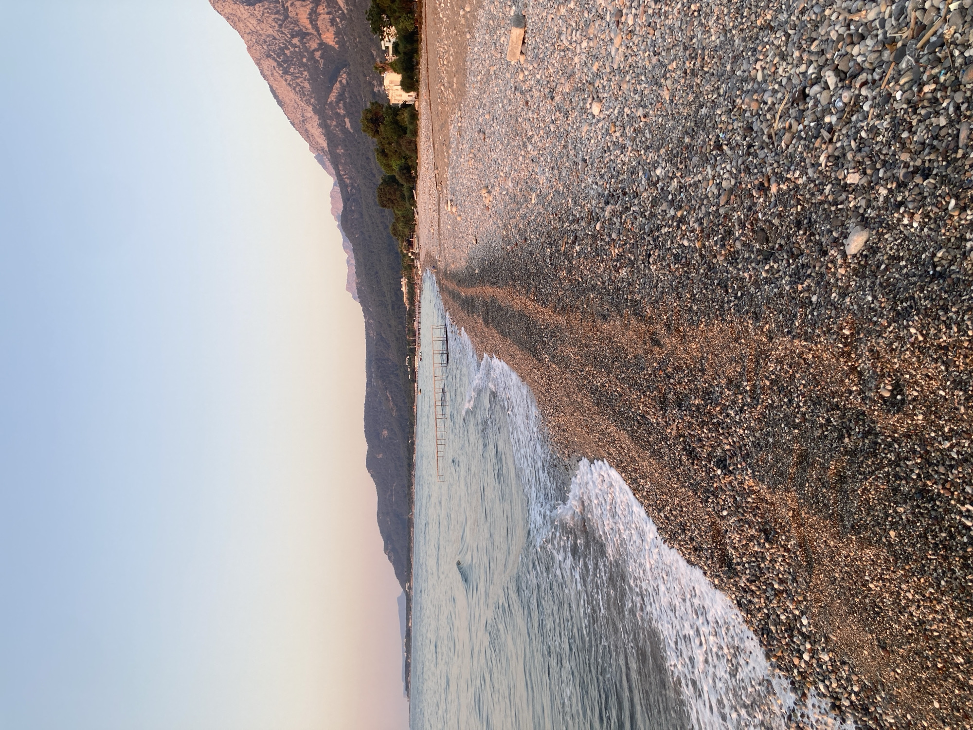 The image depicts a serene beach scene in Kemer, Turkey, at what appears to be sunset or sunrise due to the soft, warm light illuminating the landscape. The foreground is dominated by a vast expanse of pebbles that make up the beach, varying in shades of gray, brown, and white. A gentle wave, white with foam, recedes from the shore, leaving a wet sheen on the pebbles closest to the water.
The sea stretches out to the left, its surface rippled and calm, reflecting the soft hues of the sky. In the middle distance, a shallow wooden pier extends into the water, suggesting a recreational area. Along the coastline, a line of trees forms a verdant backdrop to the beach. Beyond the trees, a mountain range rises, its slopes covered in vegetation and its peaks bathed in the warm light. A few white buildings are visible nestled amongst the trees, indicating a small settlement or resort. The sky above transitions from a pale blue at the top to a soft pink and orange near the horizon, characteristic of twilight. No people are visible in the frame, contributing to the tranquil and undisturbed atmosphere of the location.