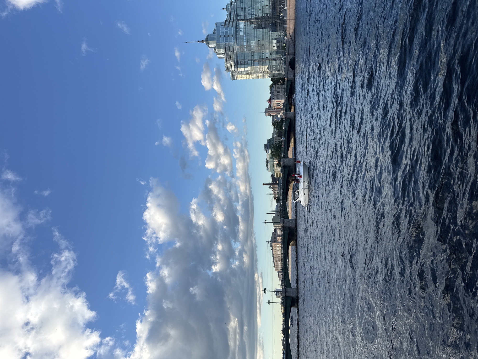 This image captures a scenic view of the Neva River in Saint Petersburg, Russia, on a cloudy but bright day. The foreground is dominated by the dark blue, rippling water of the river. In the middle ground, a white motorboat with several individuals on board is cruising, leaving a wake behind it. Further back, a long, multi-arched bridge spans the river, with lampposts and buildings lining its length. To the right of the bridge, a distinctive modern glass-covered building with a pointed spire stands out against the skyline. The background features a blend of historical and contemporary architecture, under a vibrant blue sky with scattered white clouds.
