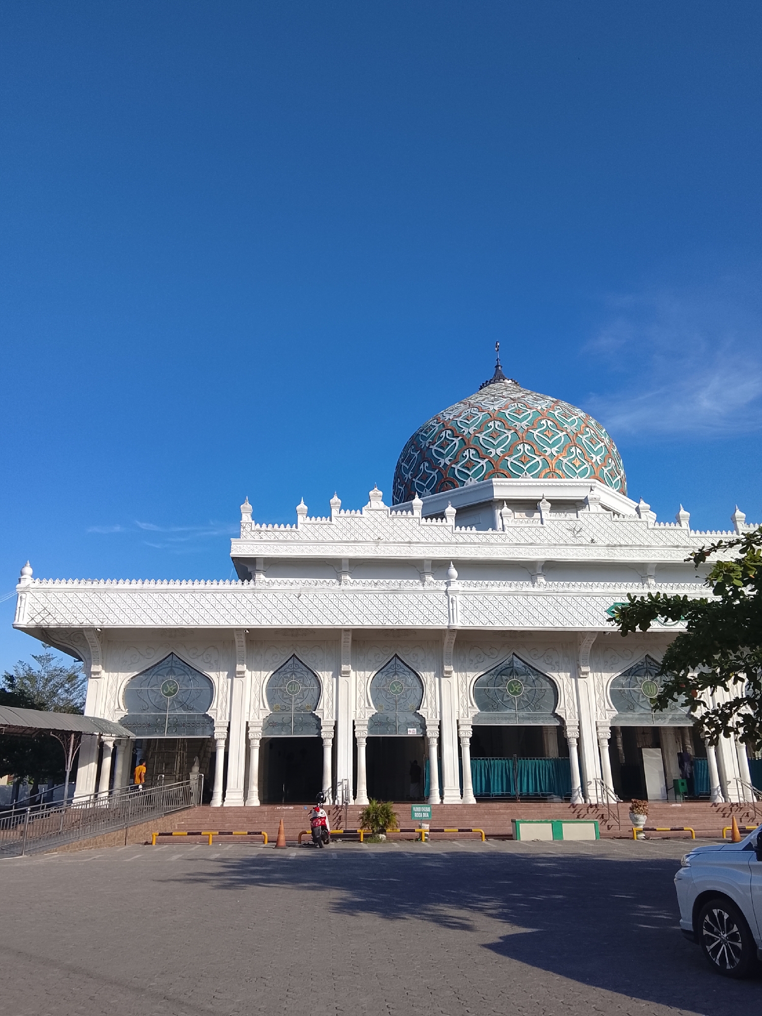 This is a low-angle, exterior, daytime, eye-level shot of the Baiturrahman Grand Mosque in Lhokseumawe, Aceh, Indonesia. The mosque features a white, intricately decorated facade with arched windows, a prominent dome adorned with geometric patterns in teal and orange, and a clear blue sky above. In the foreground, there is a paved area with yellow and orange bollards, a parked white car, and a motorcycle. A person in an orange shirt is visible walking up a ramp on the left side of the mosque entrance. The overall scene is clean and well-maintained, under bright daylight.