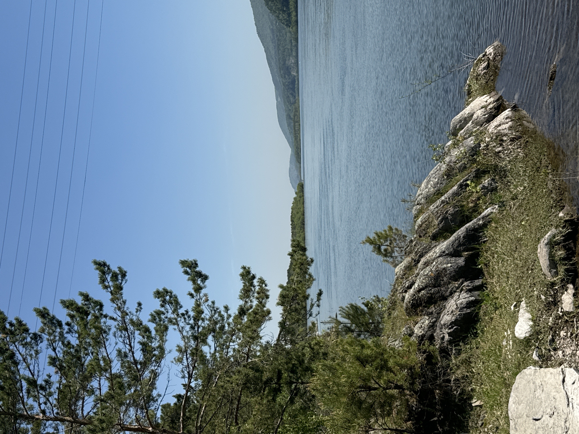 The image displays a scenic natural landscape, typical of the Krasnoyarsk region in Russia, viewed under a clear and bright sky. The photograph is oriented with the horizon running vertically, placing the sky on the left and a body of water on the right.
The left half of the frame is dominated by a vast expanse of light blue sky, indicating fair weather and bright daylight, likely midday. Several thin, dark power lines stretch diagonally across the upper portion of the sky, introducing a subtle element of human infrastructure.
In the bottom section, a dense cluster of evergreen trees, likely conifers, shows detailed branches and needles. Below and amidst these trees, and extending towards the bottom-right, are various rock formations and sparse green vegetation, creating a rugged shoreline.
The right half of the image features a wide body of water, possibly a river or lake, with noticeable ripples on its surface that suggest a gentle current or breeze. The water appears a muted blue-grey, reflecting the sky. Along the immediate right edge of the water, a prominent rocky outcrop, partially covered with greenery, juts into the water. Beyond the water, in the background, a series of forested mountains or hills recede into the distance, appearing progressively lighter and hazier.
The scene is tranquil and devoid of any human activity. No text is visible in the image. The overall impression is one of natural beauty, combining the serene expanse of water and sky with the ruggedness of the forested mountains and rocky foreground.