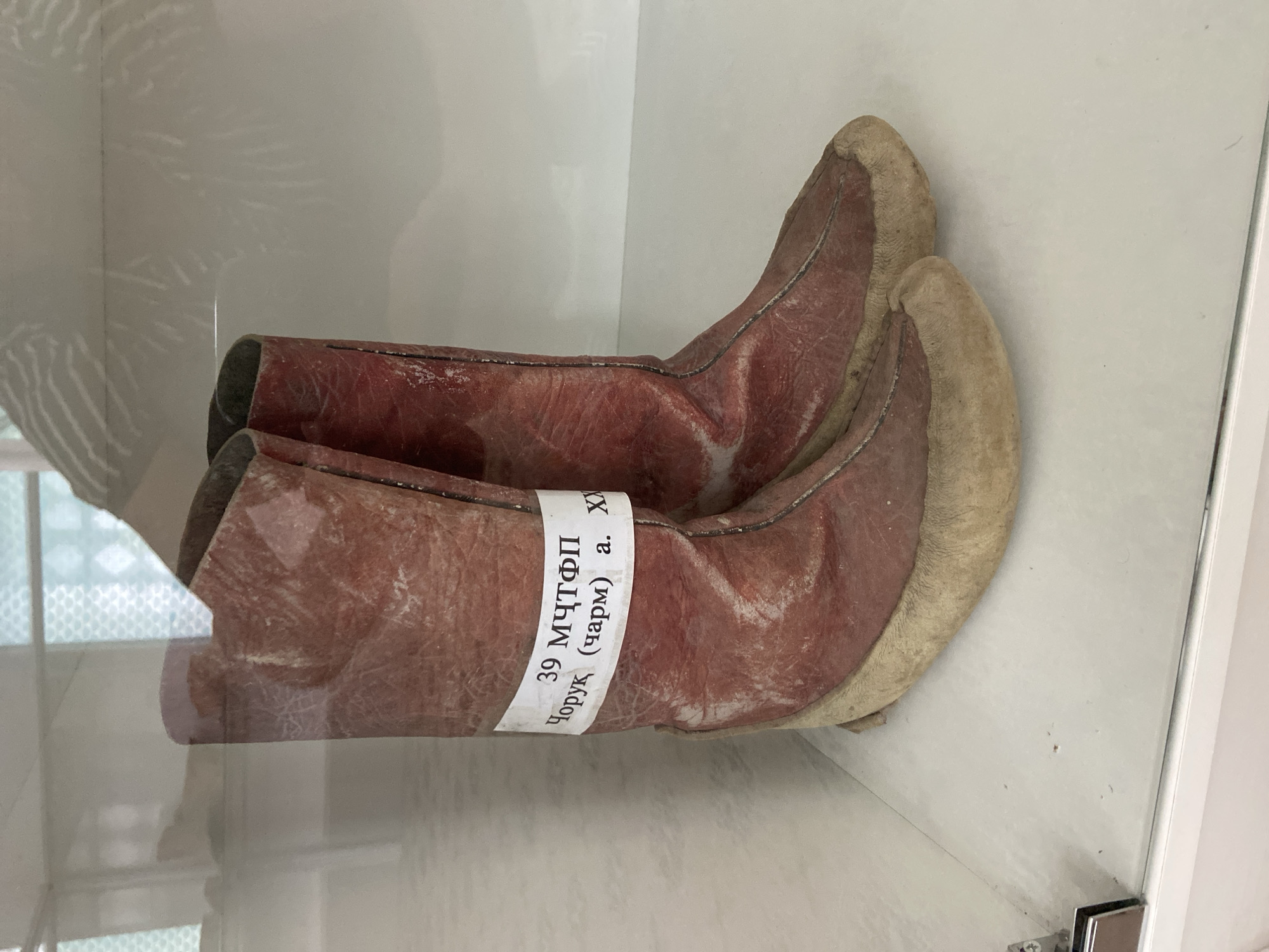 A pair of weathered, reddish-brown leather boots are displayed in a glass case. A white paper label is wrapped around the shaft of the left boot. The label contains text in a non-English script, appearing to be Cyrillic, which reads "39 МЧТФП Чopyк (чарм) а. XX". The boots have lighter-colored, possibly fur-lined, soles. They are positioned centrally within the frame, with the background of the display case appearing out of focus and reflective. The environment suggests an indoor exhibition or museum setting, possibly in Panjrud, Tajikistan, given the context provided.