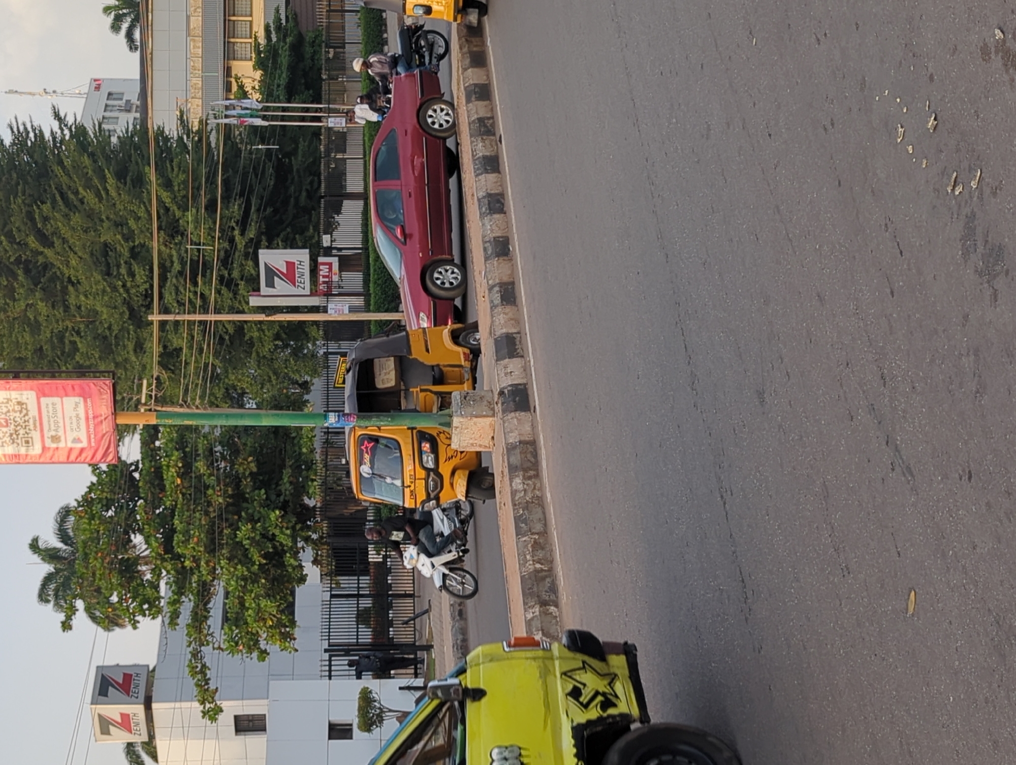 The image depicts a street scene in what is likely the city of Shaba, Nigeria, given the Zenith Bank signage in the background, a prominent bank in Nigeria. Multiple vehicles are visible, including a red sedan parked alongside the road, a yellow auto rickshaw (keke) and a green vehicle. People can be seen on a motorcycle and other unidentified people are standing on the sidewalk nearby. The street appears paved, and the environment suggests an urban setting with trees and buildings visible in the background.