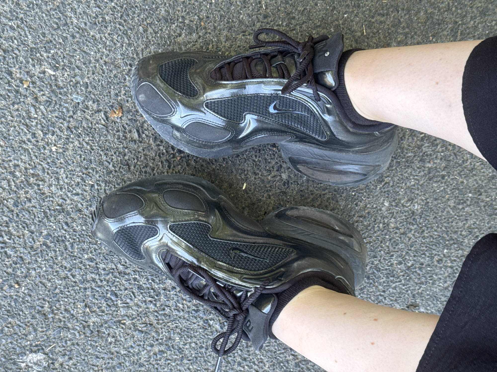 The media file features a top-down view of a person's legs wearing black Nike sneakers. The shoes have a glossy black finish with mesh detailing and black laces. The person is wearing black trousers or shorts. The scene is set outdoors on a textured grey surface, possibly asphalt or pavement. A few small flecks of debris, such as leaves and light-colored specks, are visible on the ground. The lighting suggests it is daytime, possibly sunny given the reflections on the shoes. The overall impression is casual, possibly indicating the person is walking or standing. There is no discernible text visible in the image, except for a small Nike logo on the side of the right shoe. The location context is specified as Brussels, Belgium.