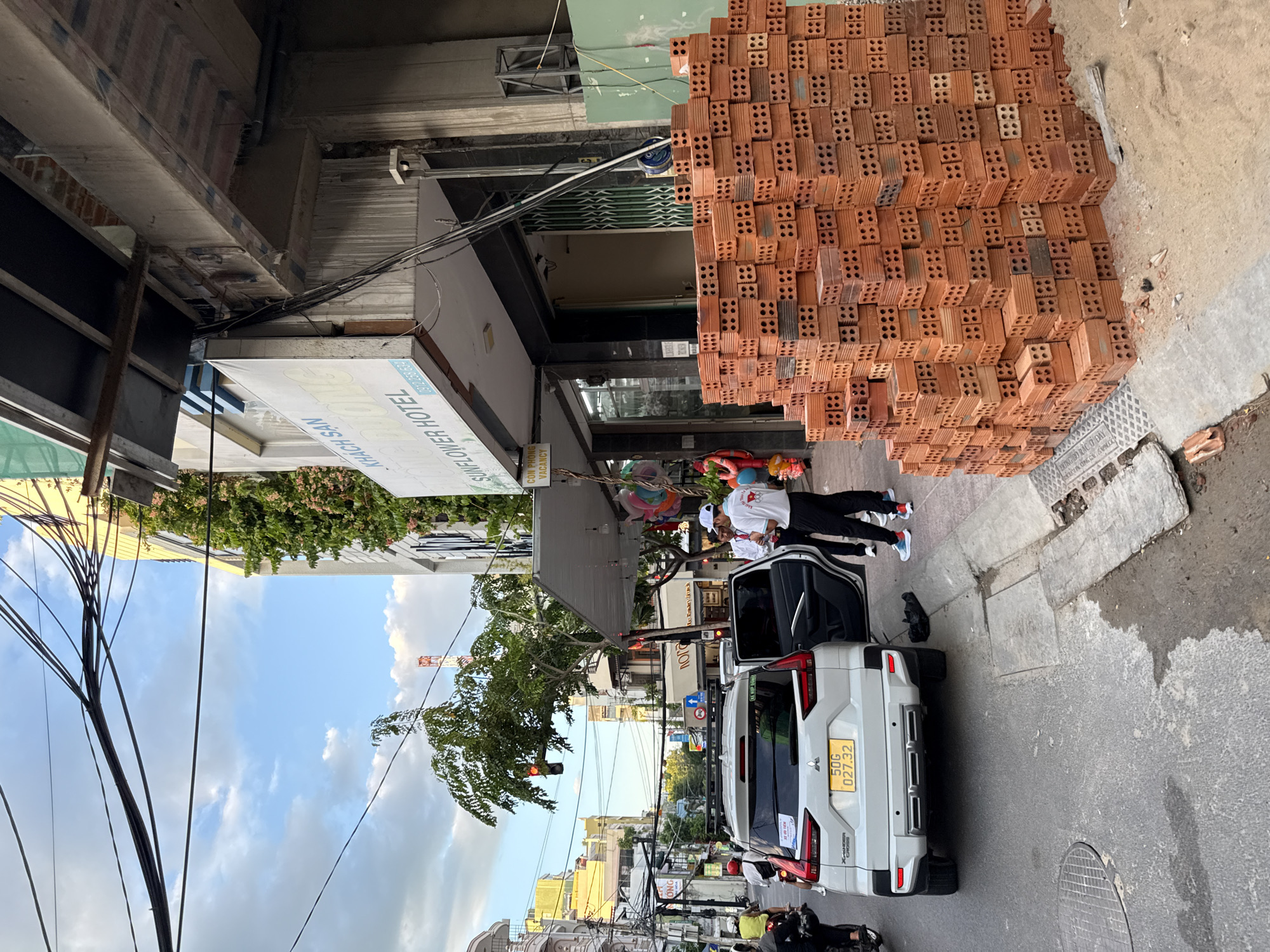 In Nha Trang, Vietnam, a street scene unfolds under a cloudy sky with overhead power lines. A white Mitsubishi Xpander Cross car, license plate 50G 02732, is parked on the side of the road with its rear passenger door open. Two men are standing near the open car door. One man, wearing a white polo shirt with red trim and a white cap, stands with his hands behind his back. The second man, wearing a white collared shirt and black pants, is partially obscured by the first. In front of a building, a large stack of orange bricks is piled on the sidewalk, suggesting construction or repair work. The building has a sign for "SUNFLOWER HOTEL" and a smaller sign indicating "CON PHONG VACANCY." Green foliage is visible climbing the side of the building. The street is paved with asphalt and concrete, showing some wear and tear. Several motorcycles are seen in the background, indicating typical city traffic.