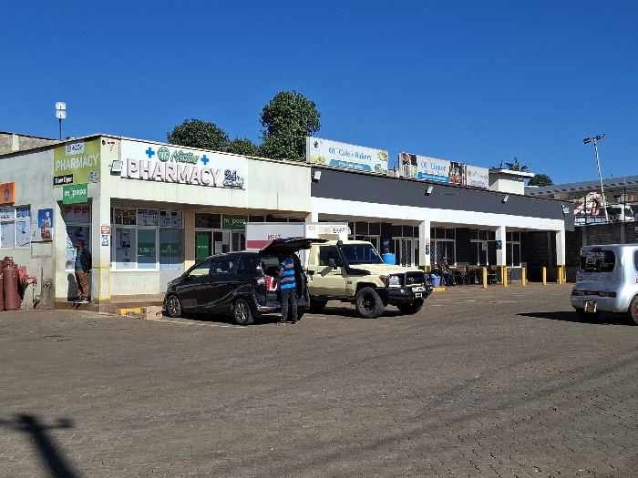 The image shows a street scene in Gitaru, Kenya (latitude: -1.231, longitude: 36.668). There is a pharmacy on the left side of the image, with a sign that says "The Master Pharmacy 24/7". Next to the pharmacy is a larger building with a sign that says "CC Cafe & Bakery" and another sign that reads "CC Liquor". There are two cars parked in front of the buildings, a black car with its trunk open and a beige pickup truck. A man wearing a blue shirt is loading items into the black car. The buildings are situated on a paved street with a brick-like pattern. The sky is clear blue. There are trees in the background. There is a red fire hydrant near the corner of the pharmacy building. There are yellow poles in the street. There is a pedestrian walking on the sidewalk in front of the pharmacy. The street appears to be fairly quiet, with only a few people visible.