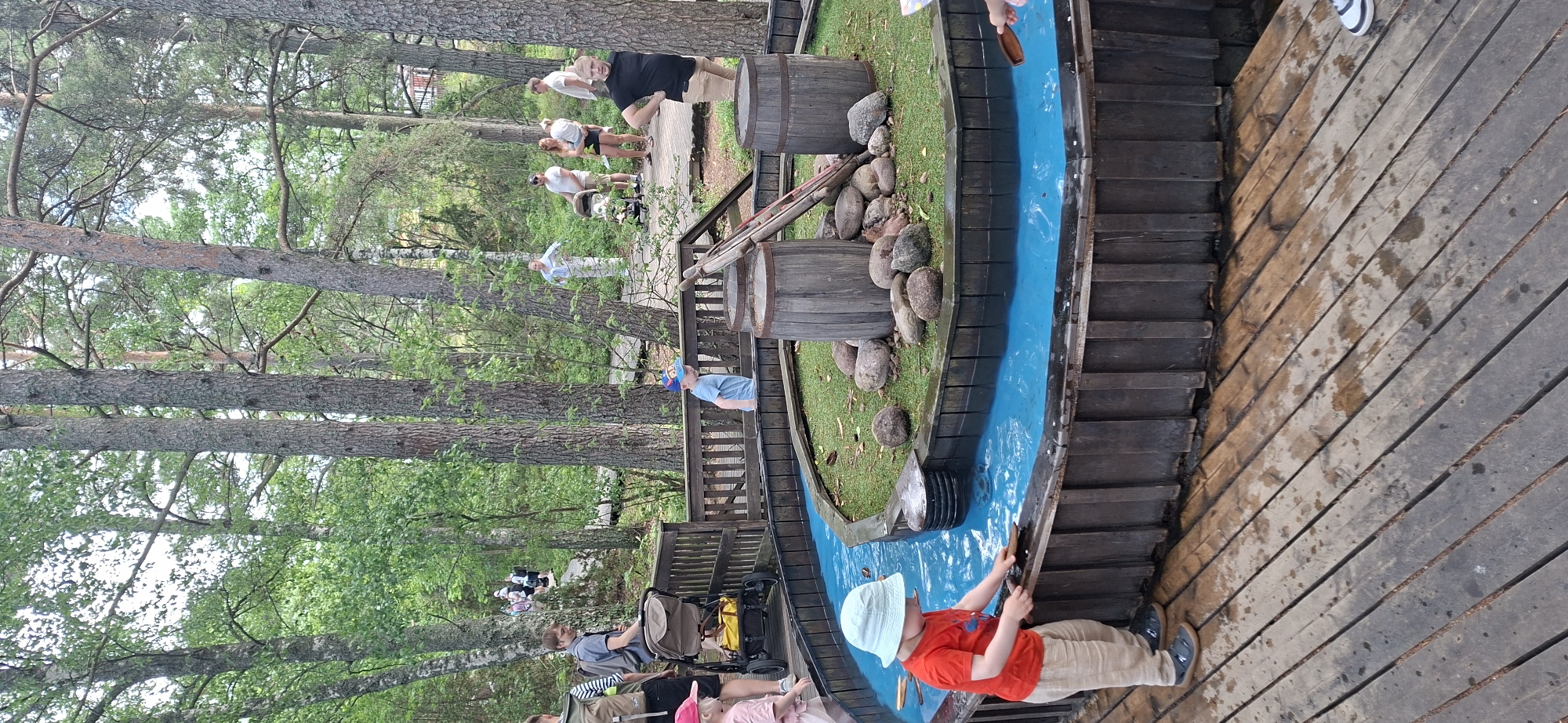 In Naantali, Finland, at an outdoor attraction, a young boy in a red shirt, beige pants, and a light blue hat stands on a wooden deck, playing in a circular water feature with blue water. He holds a wooden scoop in his hands, engaging with the water. The water feature has wooden planking and is surrounded by grass and large stones. Two wooden barrels and a red ladder-like structure are present within the feature. In the background, several adults and children are visible on wooden walkways amidst a forest setting with tall trees. One adult pushes a stroller. The overall environment suggests a family-friendly recreational area.