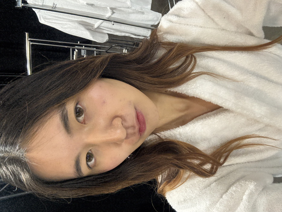 A young woman is captured in a close-up, chest-up shot, looking directly into the camera. She has long, dark brown hair with lighter auburn highlights, which falls over her shoulders and partially frames her face. Her head is tilted slightly to her left, creating a candid pose. She is wearing a white, textured bathrobe, suggesting a relaxed or post-bath setting. Her complexion is fair, with a natural, rosy flush on her cheeks, and her lips are colored with a pinkish-red tint. Her eyes are dark brown and meet the viewer's gaze with a calm, neutral expression.
In the background, out of focus, there is a metallic rack, possibly a towel or clothes rack, draped with several white towels or linens. The setting appears to be indoors, likely a bathroom, hotel room, or domestic environment, given the presence of the bathrobe and towels. The lighting is soft and even, suggesting indoor artificial light or diffuse natural light. There is no visible text in the image. The overall impression is one of relaxed intimacy.