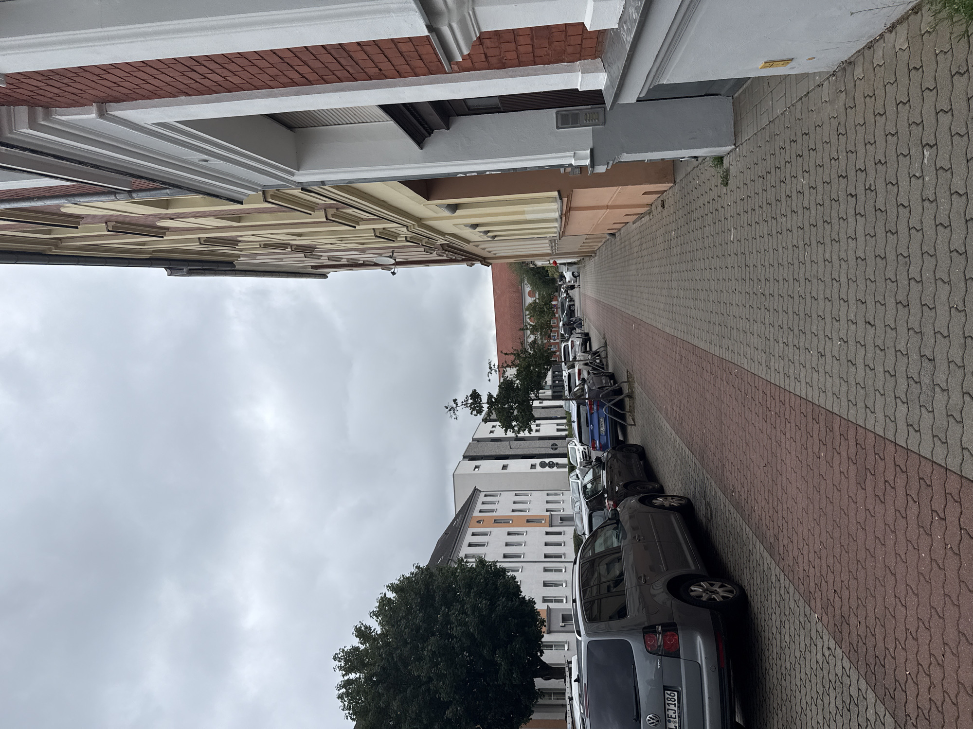 This image depicts a street scene in Rathenow, Germany, on a cloudy day. The perspective is from a low angle, looking along a sidewalk lined with parked cars and buildings. The sidewalk is paved with interlocking stones in shades of gray and red.
On the left side of the street, a row of buildings is visible. The closest building features ornate architectural details, including red brickwork and white decorative trim. Further down the street, modern apartment buildings with numerous windows are prominent. A large, mature tree with dark green foliage is situated in front of one of these buildings.
The right side of the street is occupied by parked vehicles. Several cars are visible, including a silver van or SUV in the foreground with a license plate that reads "P-EJ 386". Behind it, other cars of various colors are parked along the curb.
The sky is overcast, suggesting a cloudy or possibly impending rain. The time of day is unclear due to the lack of direct sunlight. The overall atmosphere is one of a quiet residential street. No people are clearly visible in the frame.