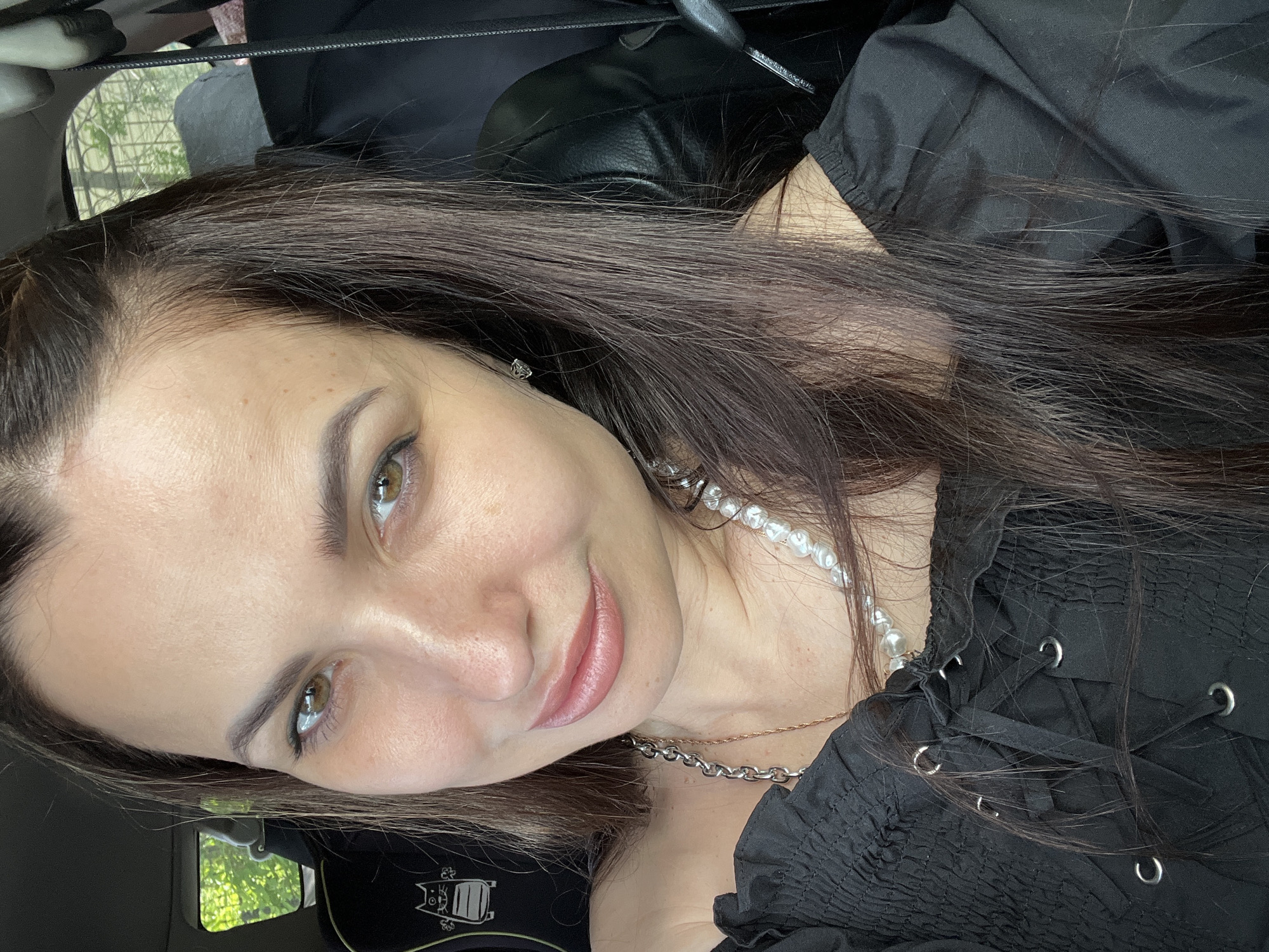 A close-up, portrait-style selfie shows a woman with long, dark brown hair and a gentle, soft smile, positioned in what appears to be a car interior. The original image is rotated counter-clockwise, with the woman's head tilted.
The subject has a light complexion with visible freckles across her forehead and nose. She has striking hazel eyes that are looking directly at the camera. Her eyebrows are well-defined, and her lips have a natural, subtle pink hue, possibly enhanced with a tint or permanent makeup. She is wearing a black top with a shirred or ruched texture, featuring a lacing detail with silver-toned eyelets on the shoulder/chest area. She accessorizes with a white pearl necklace, a silver-toned chain necklace layered beneath it, and a small silver stud earring visible on her left earlobe.
The background details suggest an automotive setting, with a black seat or headrest visible behind her. Through a car window, blurred green foliage can be seen, indicating an outdoor, daytime environment. A window shade or visor is also partially visible above the foliage. On what appears to be a headrest cover in the lower left of the frame (when viewed in the original orientation), there is a black fabric with a white, stylized graphic resembling a cat's face with ears. Any accompanying text below the graphic is too indistinct to be read clearly.
The woman's expression is calm and pleasant, conveying a relaxed and friendly mood. The lighting is bright and natural, consistent with daytime. No specific activity is depicted other than posing for the photograph.