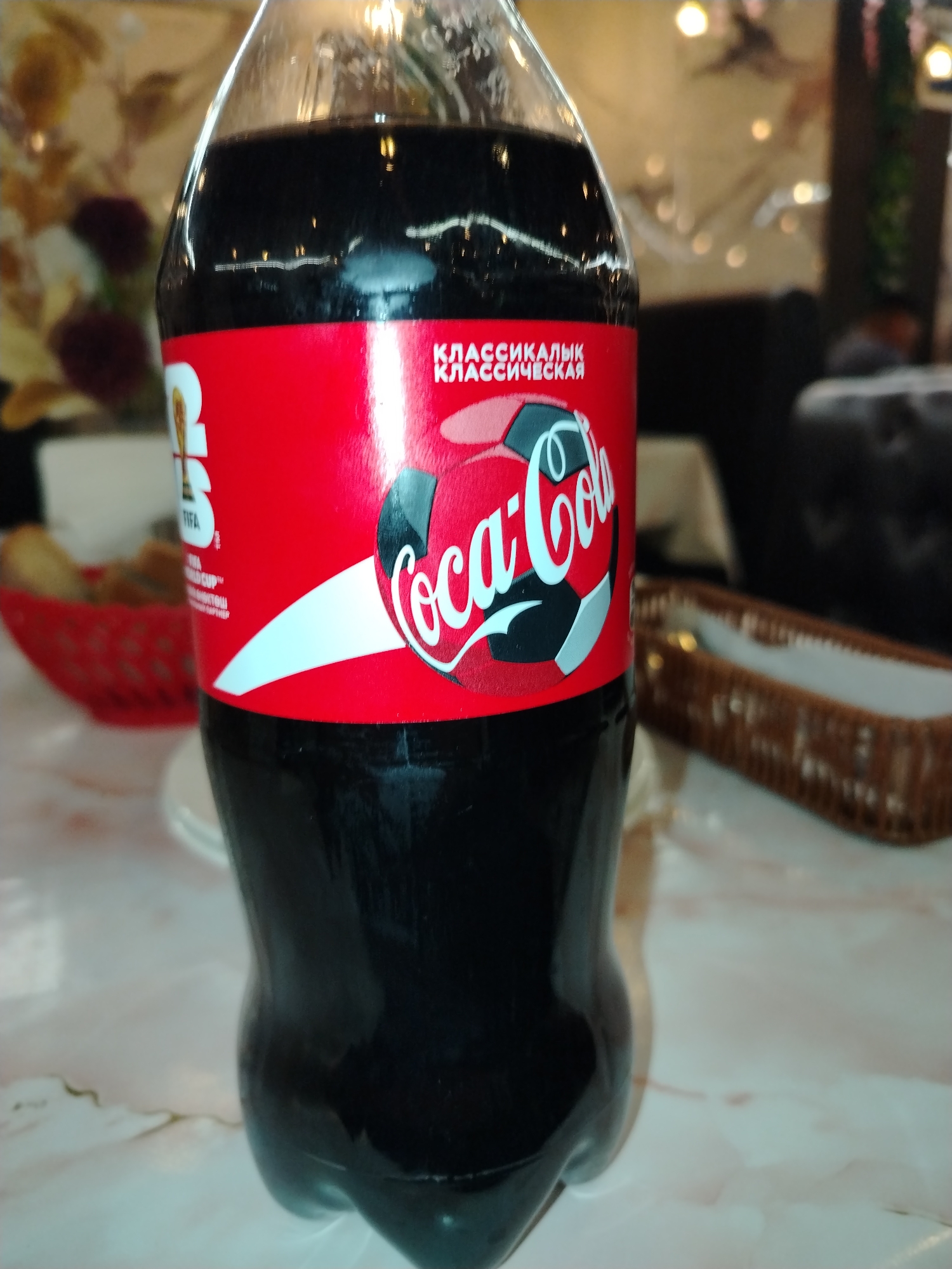 A close-up, eye-level shot captures a Coca-Cola bottle positioned prominently in the foreground. The bottle, filled with a dark liquid, is the primary subject, with its red label featuring the iconic Coca-Cola logo and a soccer ball design. To the left of the bottle, a red woven basket containing food items is partially visible. In the background, a blurred image of a dining setting is discernible, with hints of tables and seating. The overall lighting suggests an indoor environment, possibly a restaurant or cafe. The image does not provide any specific indicators of its location in Bishkek City, Kyrgyzstan.