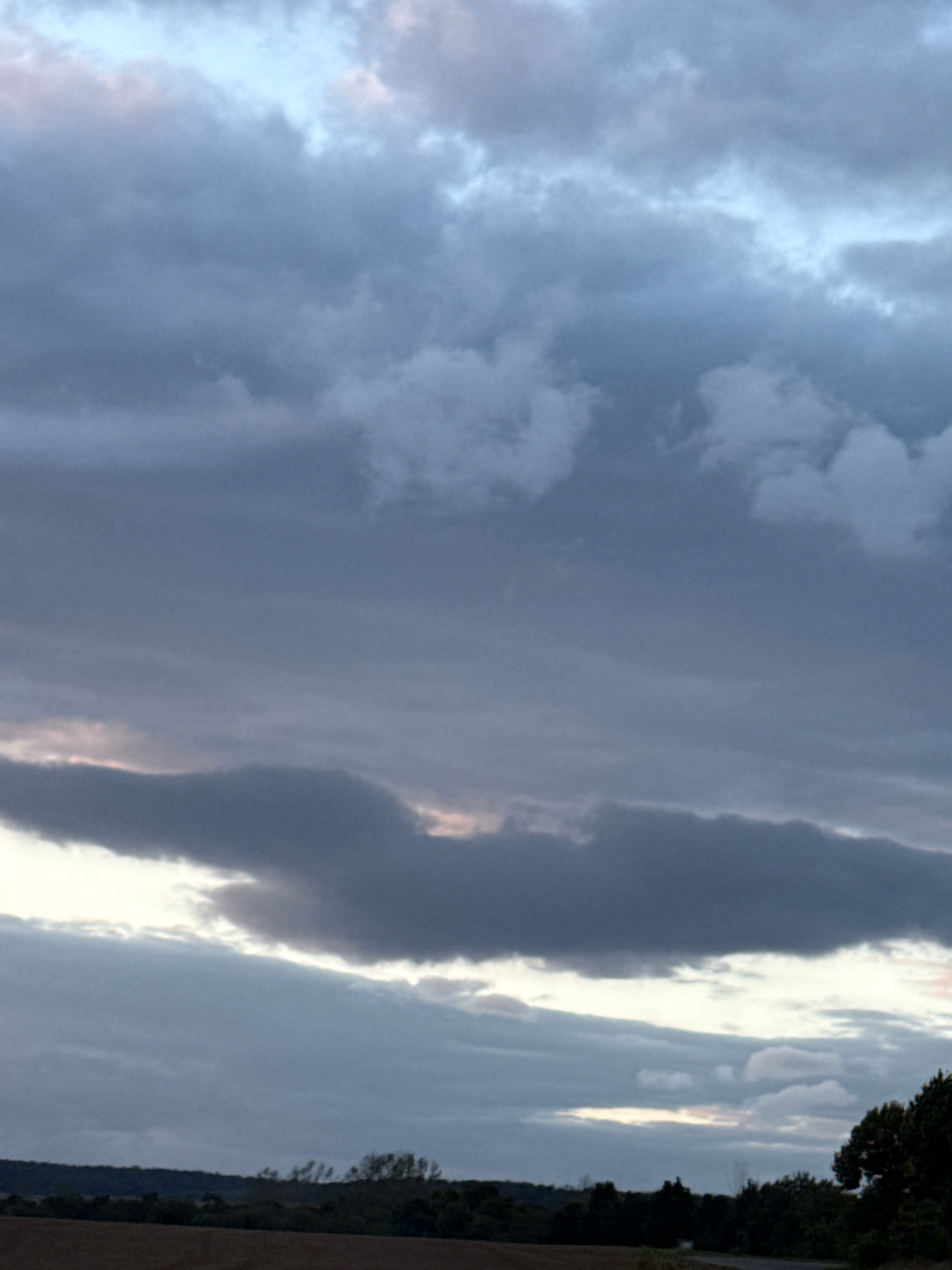 This image shows a cloudy sky over a rural landscape. The sky is filled with various shades of gray and blue clouds, with some hints of pink and yellow peeking through at the horizon. Below the sky, a dark line of trees and vegetation stretches across the middle ground. The foreground appears to be a field, possibly agricultural land. The overall impression is of an overcast day in a rural setting near Greußen, Germany.